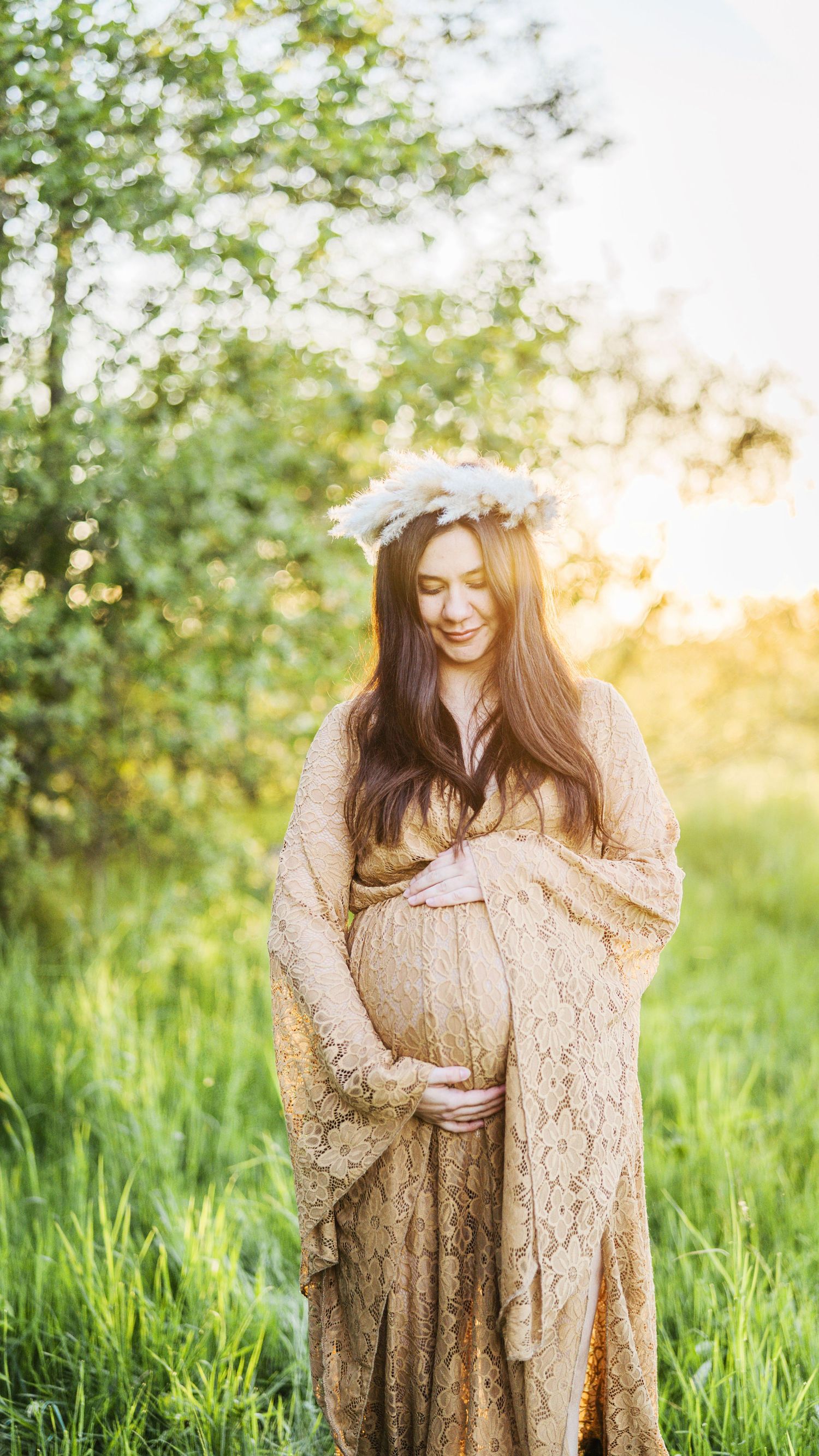 Pregnant woman in golden lace gown and white floral crown standing in sunlit green garden.