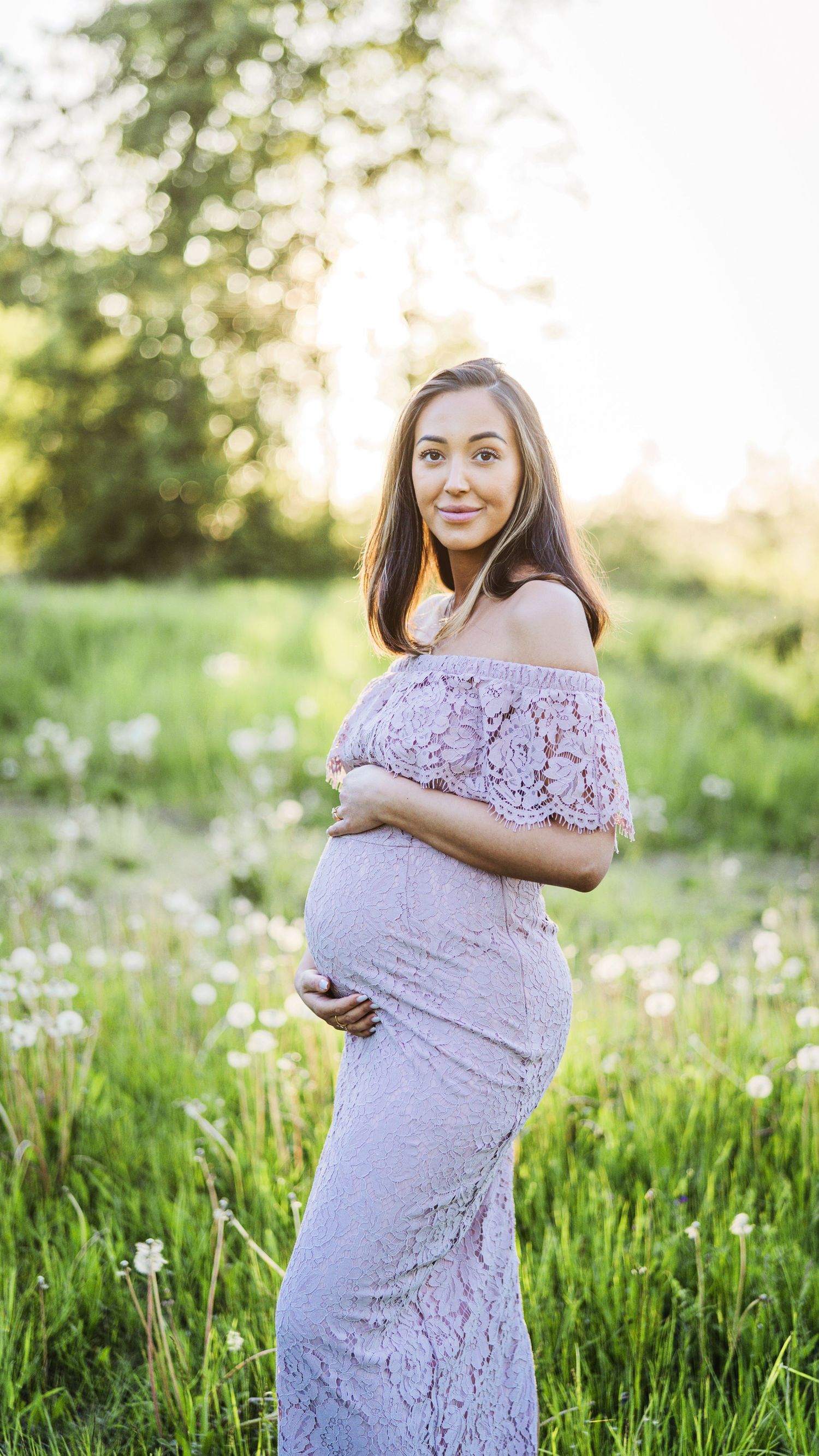 Pregnant woman in lavender lace off-shoulder gown smiles while holding bump in sunny field.