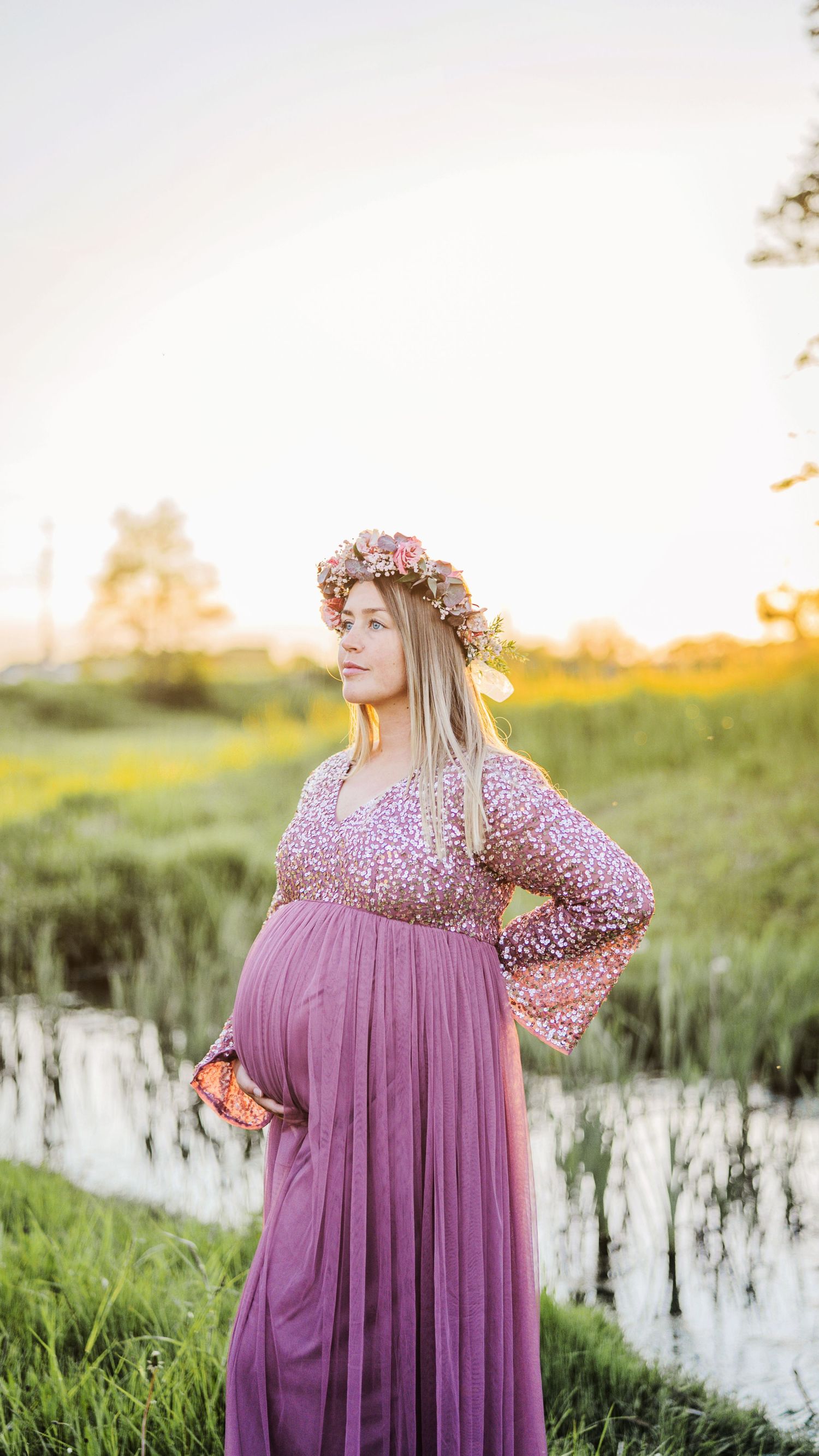 Pregnant woman in purple sequin gown and floral crown poses confidently near pond at sunset.