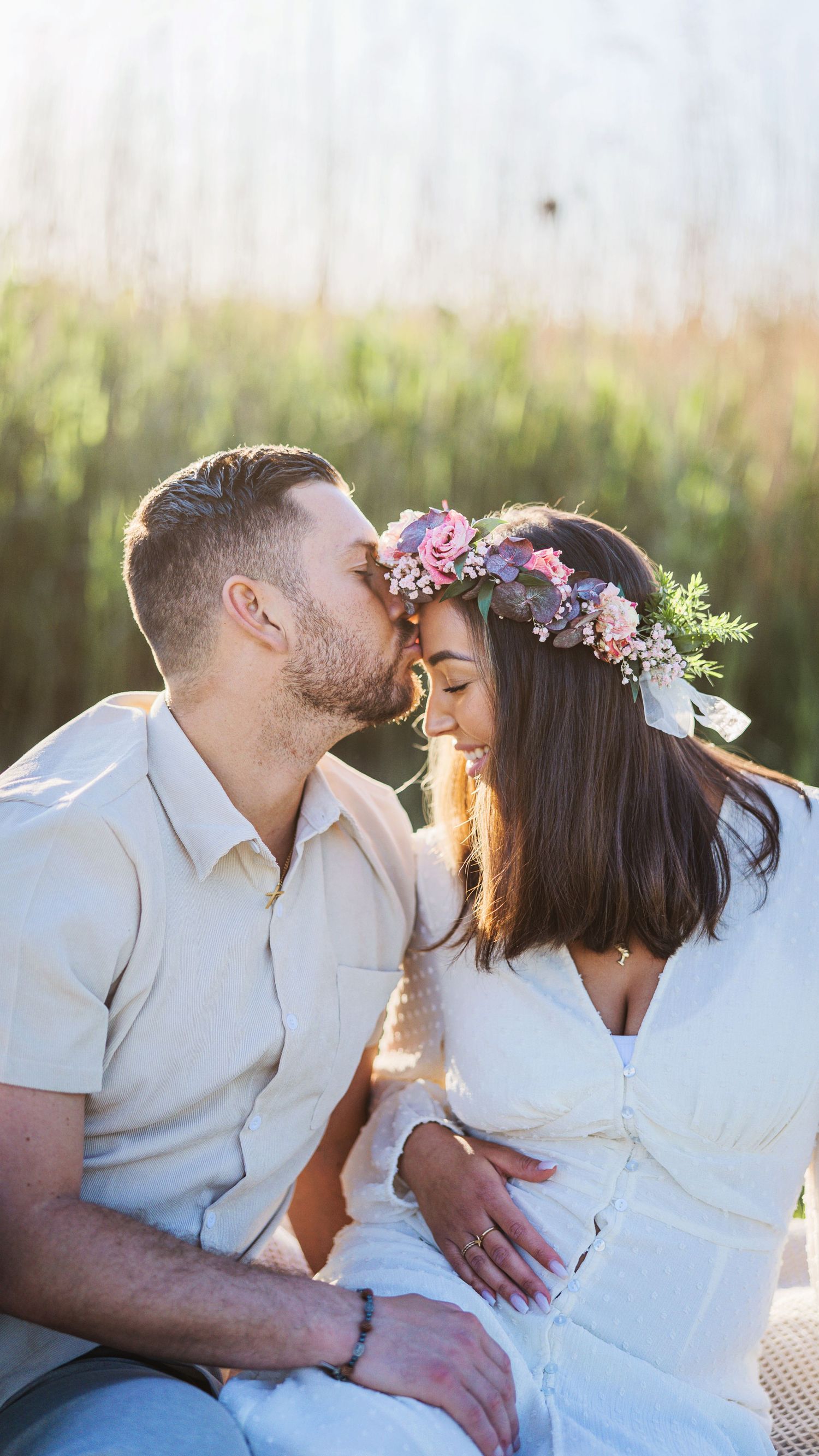 Couple sharing a tender kiss during outdoor maternity photoshoot, woman wearing floral crown, golden hour lighting.