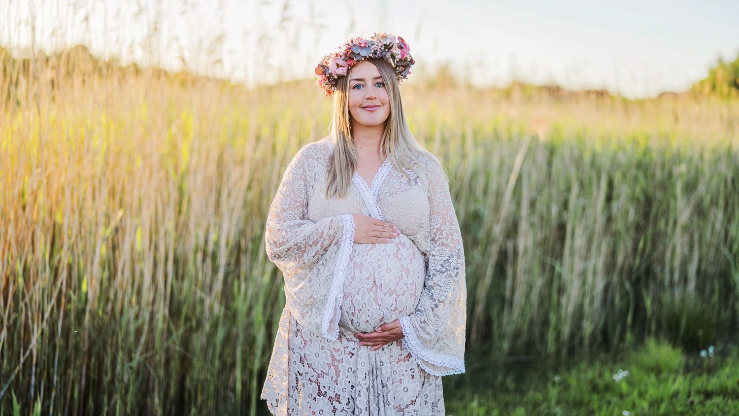 Pregnant woman in white lace dress and floral crown cradles bump near tall grass at sunset.