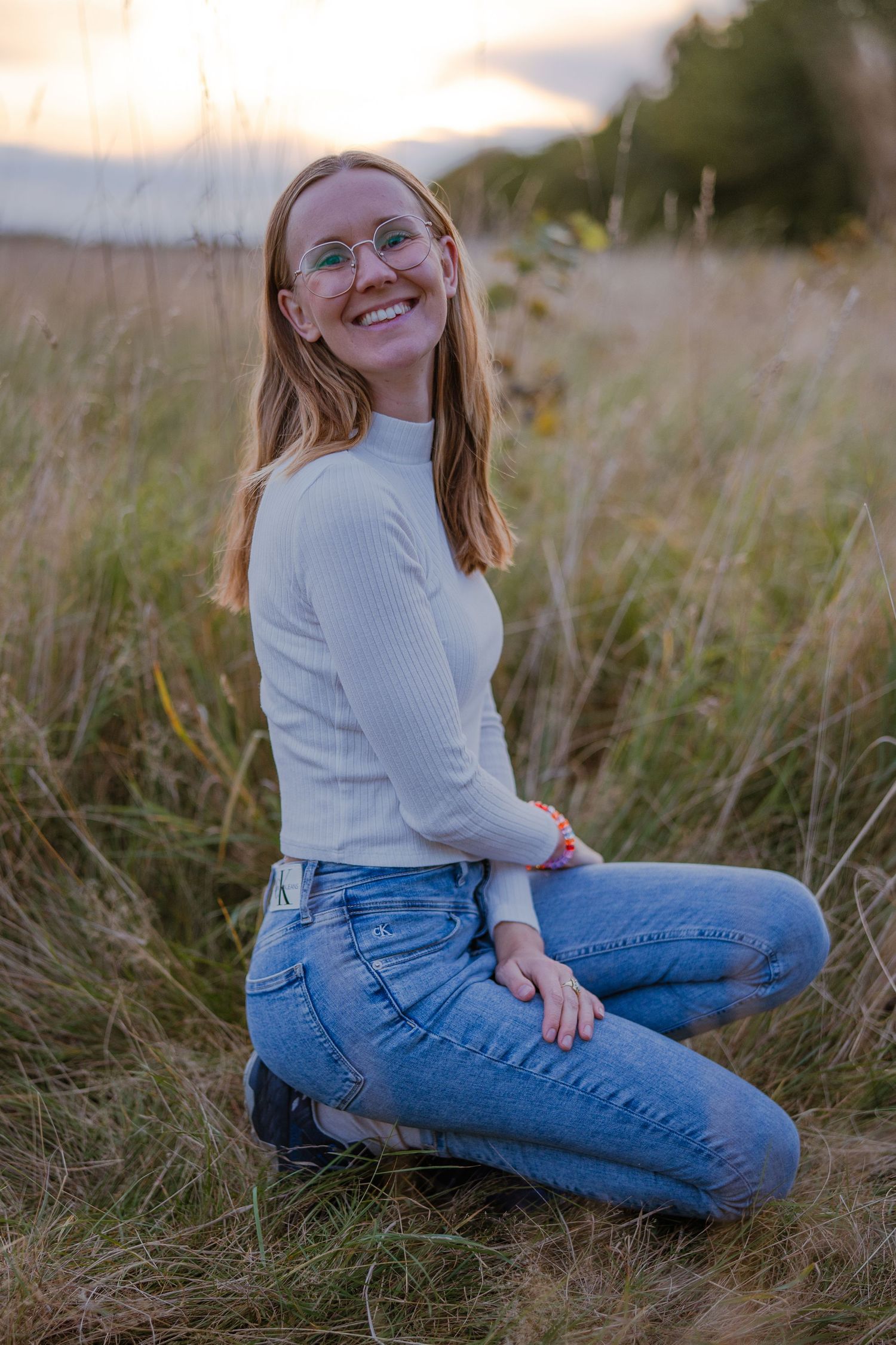 Smiling redhead woman with glasses sitting in a golden field at sunset wearing white top and blue jeans.