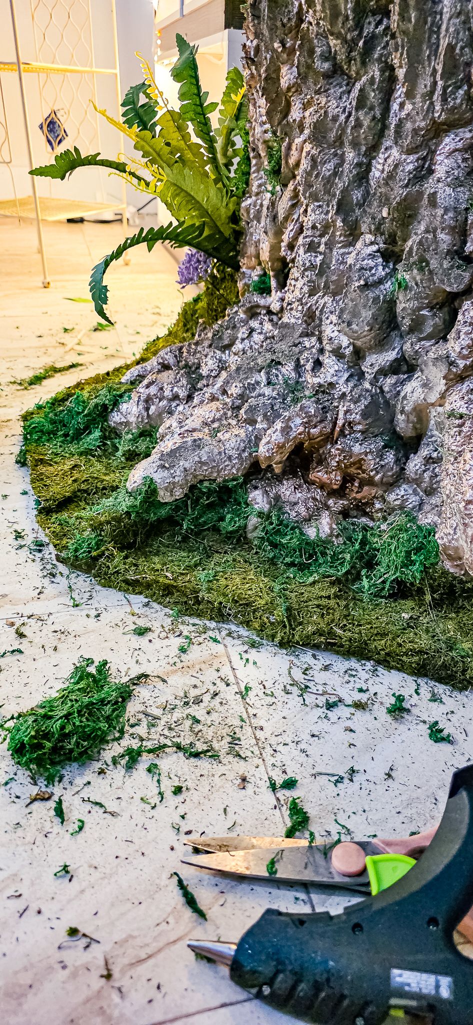 Artificial tree base with green moss, ferns, and scissors on white floor during decorative installation.