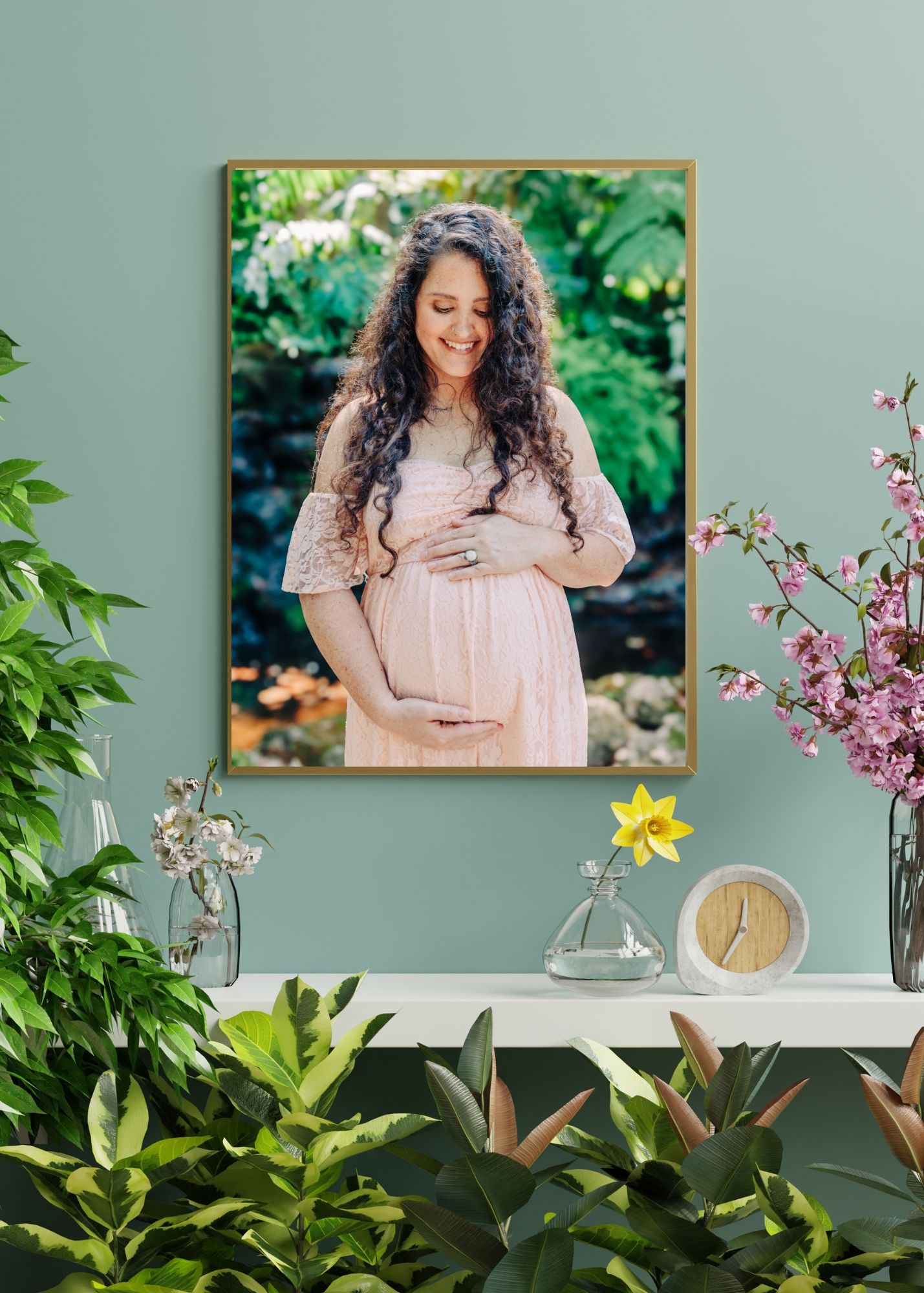 A pink-themed gallery wall and shelf decorated with maternity photos, floral accents, and greenery creates a soft nursery setting.