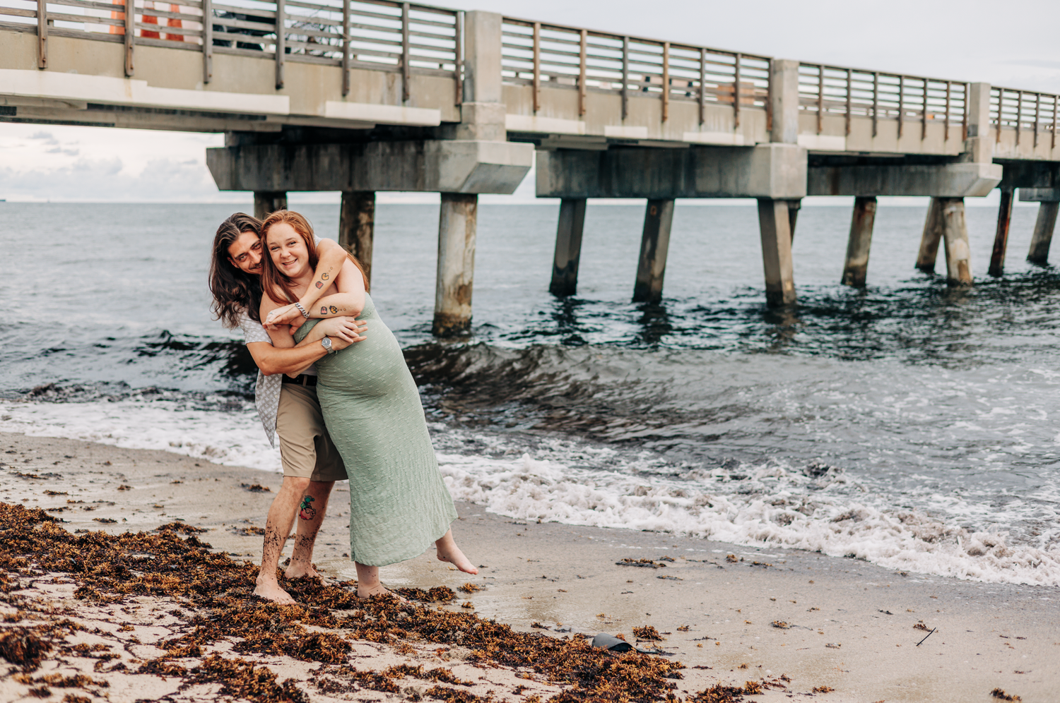Maternity photoshoot Lake Worth of an expecting couple in a sage green dress beneath the Lake Worth Beach pier.