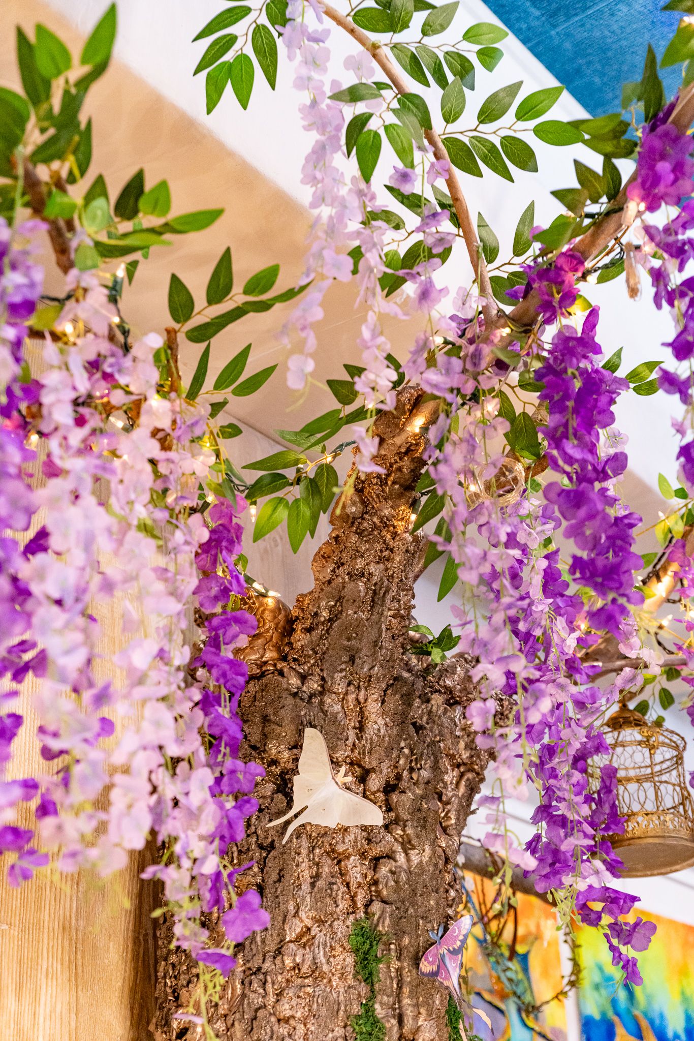 Decorative wisteria tree with cascading purple flowers, green leaves, and a white butterfly ornament in an indoor setting.