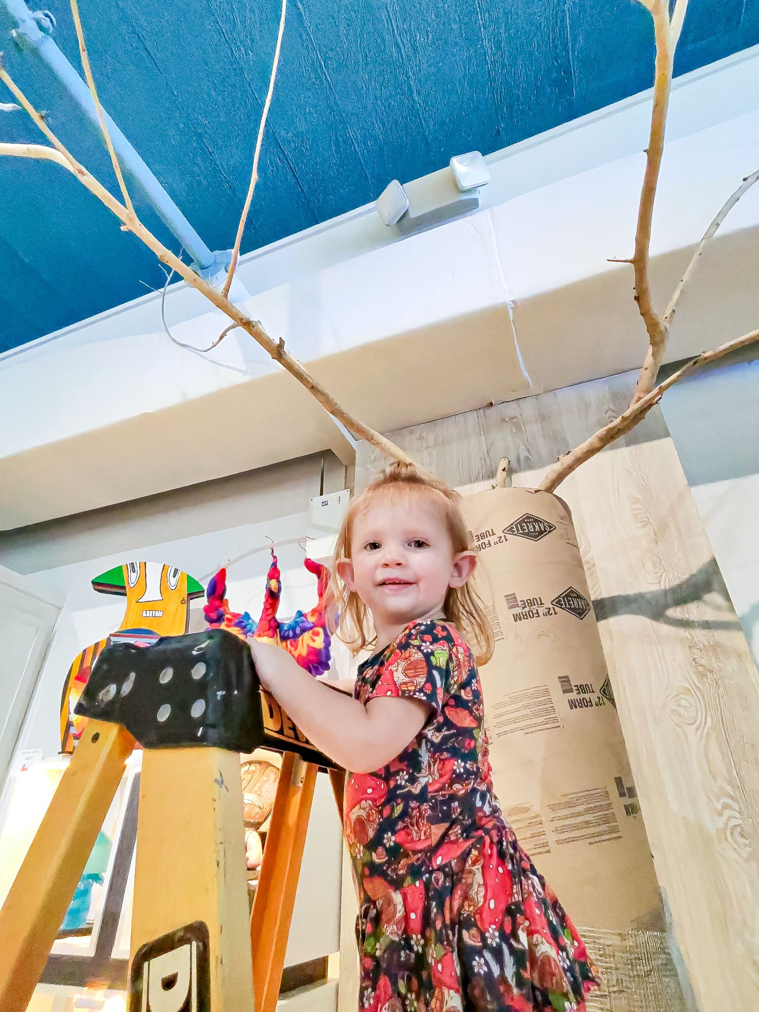 Young girl in floral dress smiling beside a large cardboard tube and bare branch art installation indoors.
