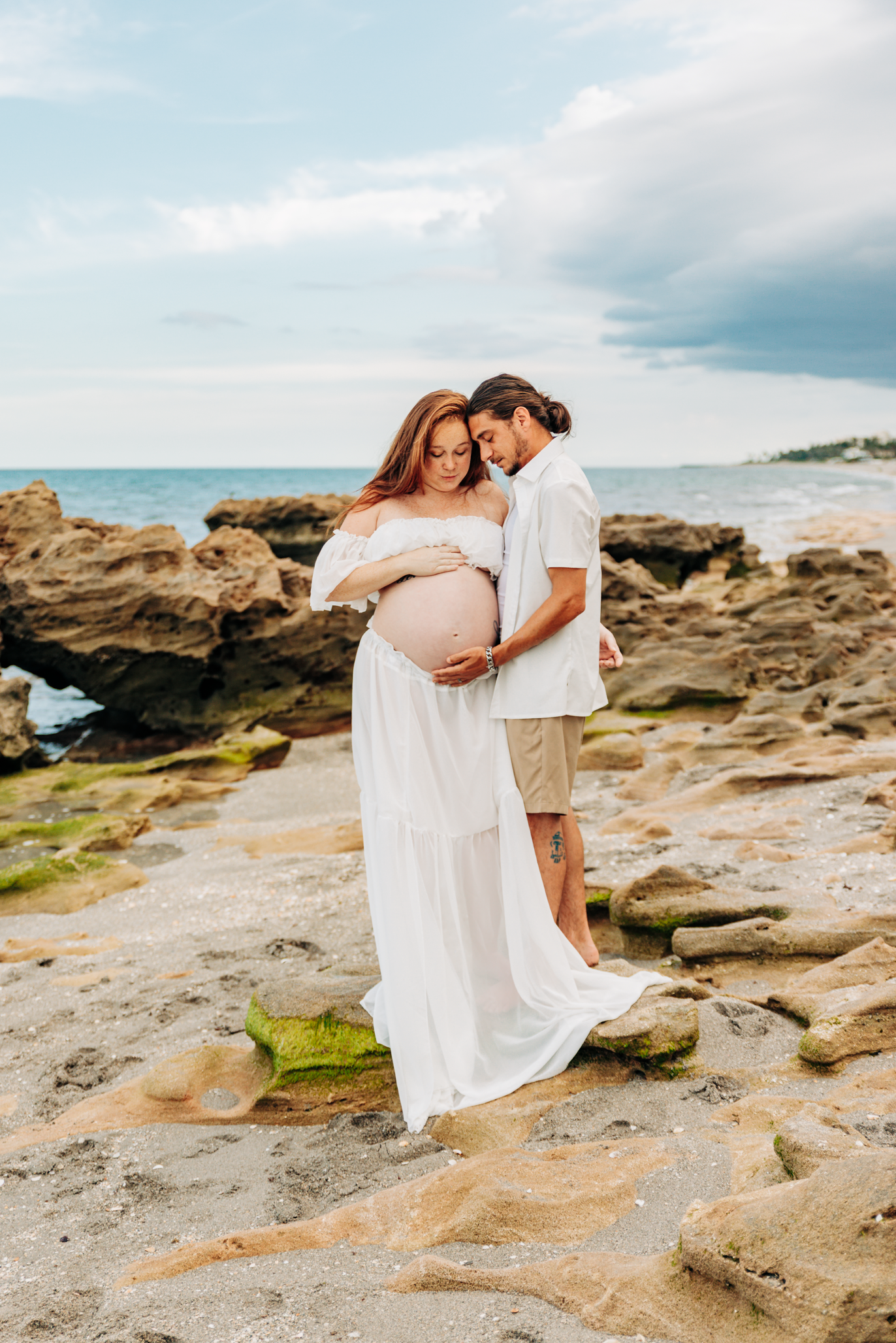 Maternity photographer Jupiter FL portrait of an expecting couple embracing on the rocky shoreline at Coral Cove Beach.