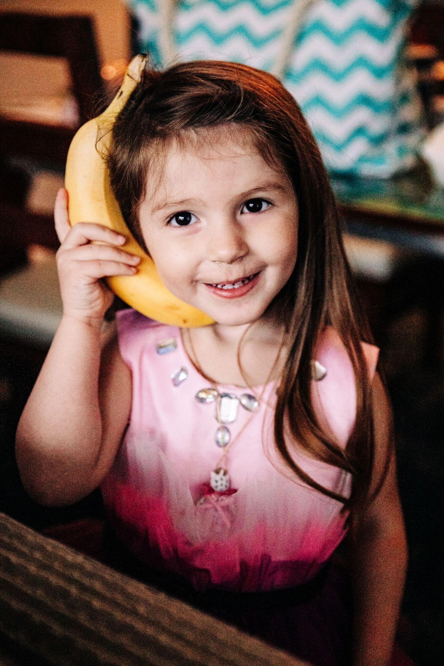 A young child wearing a pink dress holds a banana up to their ear like a telephone with a cheerful expression.