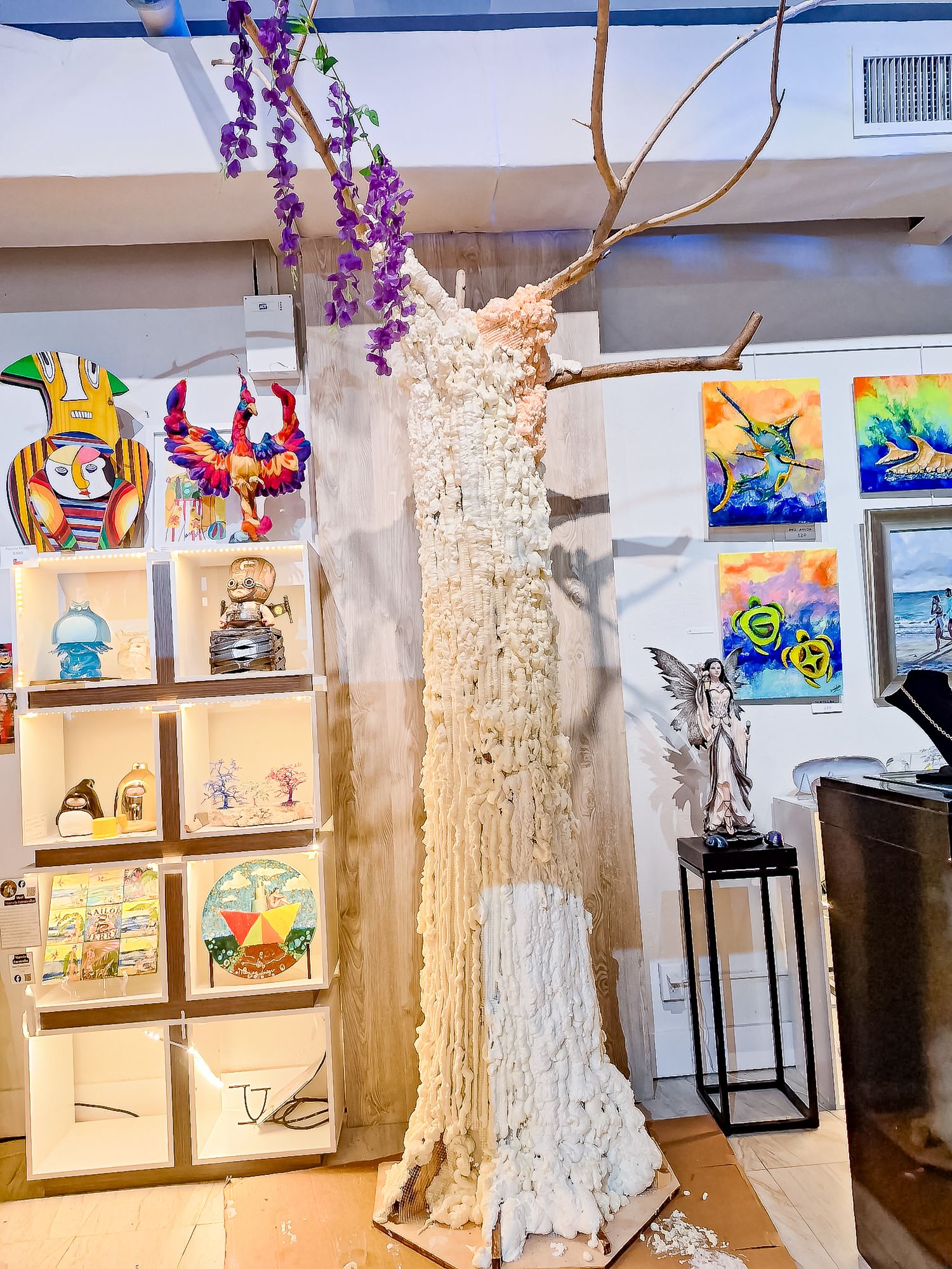 Art gallery displaying a tall textured white tree sculpture with purple flowers, colorful paintings, and a fairy figurine.