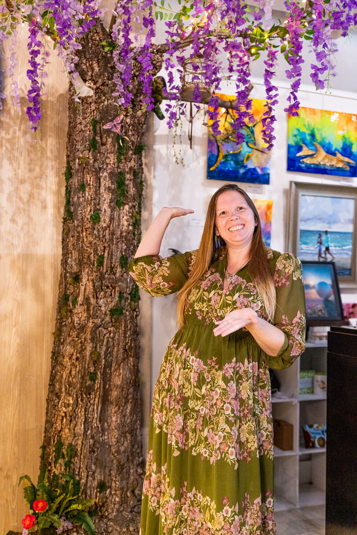 Kelsey in floral green dress posing beside decorative tree with purple wisteria flowers in an art gallery.