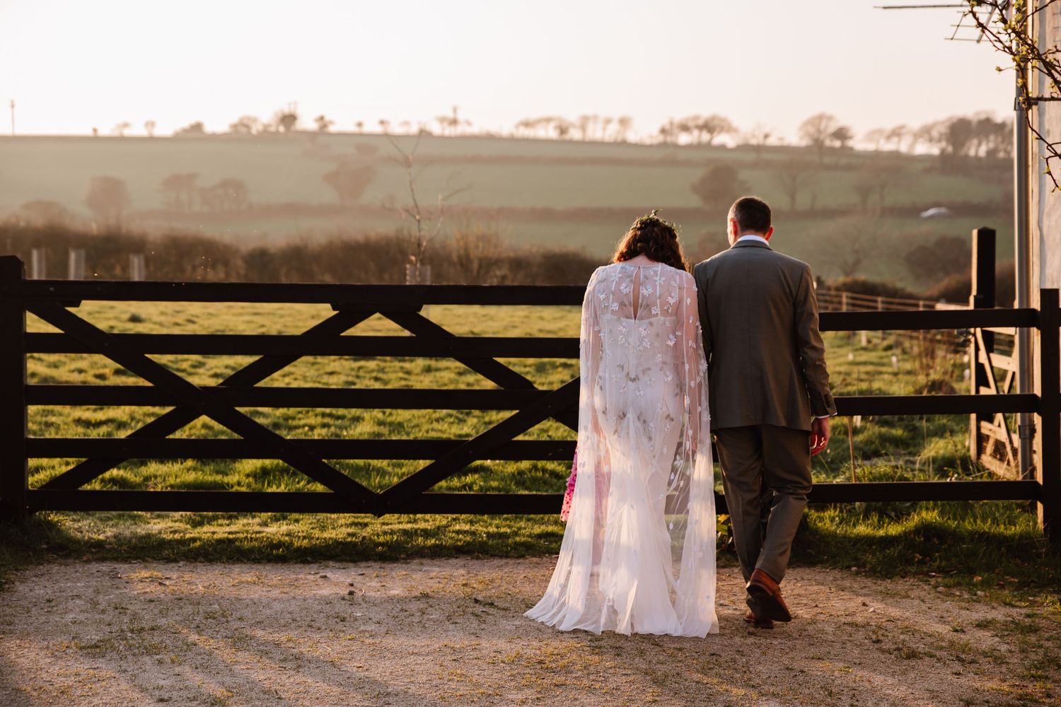 Bride and groom walking hand in hand toward a rustic wooden gate at golden hour in a countryside setting. The Cornish Place, Wood, Cornwall