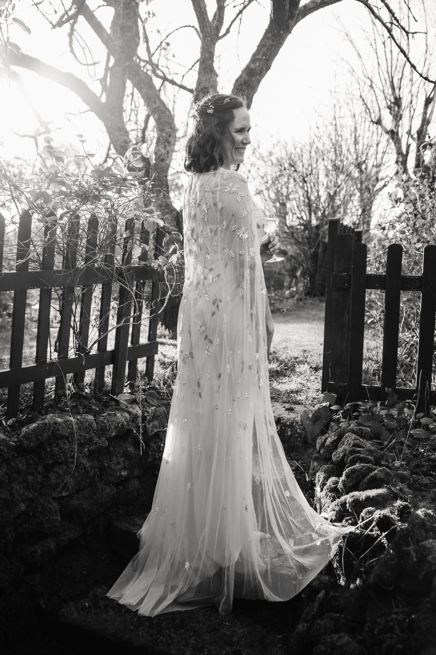 Black and white photo of a bride in a flowing white gown with cape, standing by a rustic fence in a garden. The Cornish Place, Wood shed