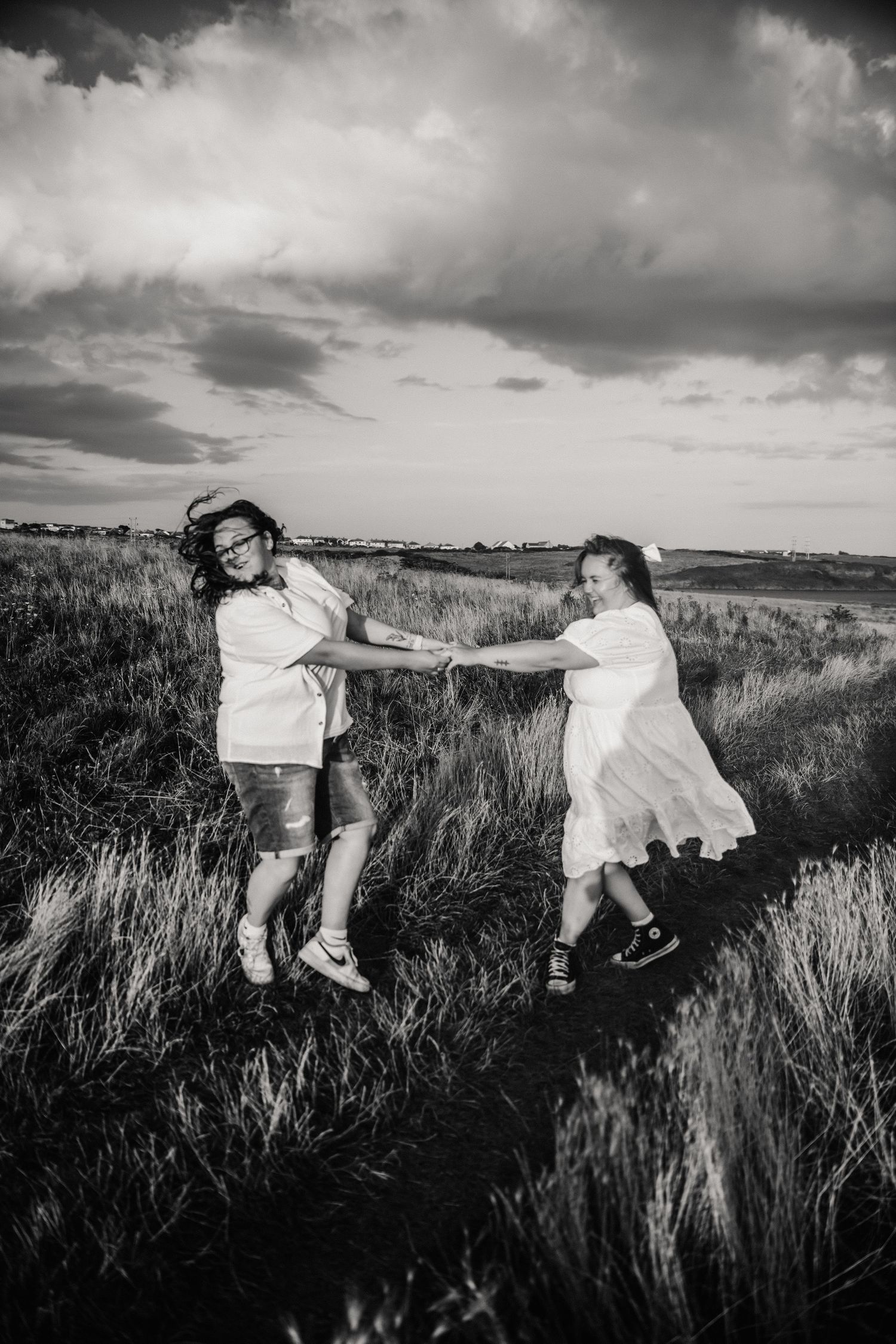 Two girls in white dresses hold hands and spin joyfully in a grassy field under a dramatic cloudy sky.