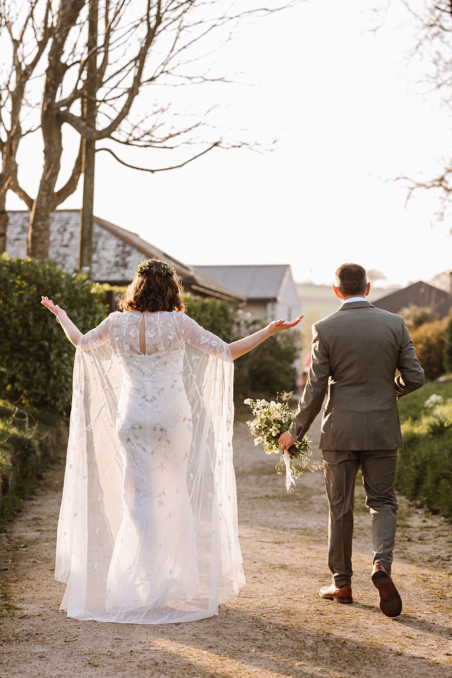 Bride in lace cape dress and groom in gray suit walk hand-in-hand down a sunlit path at their outdoor wedding at The Cornish Place wedding