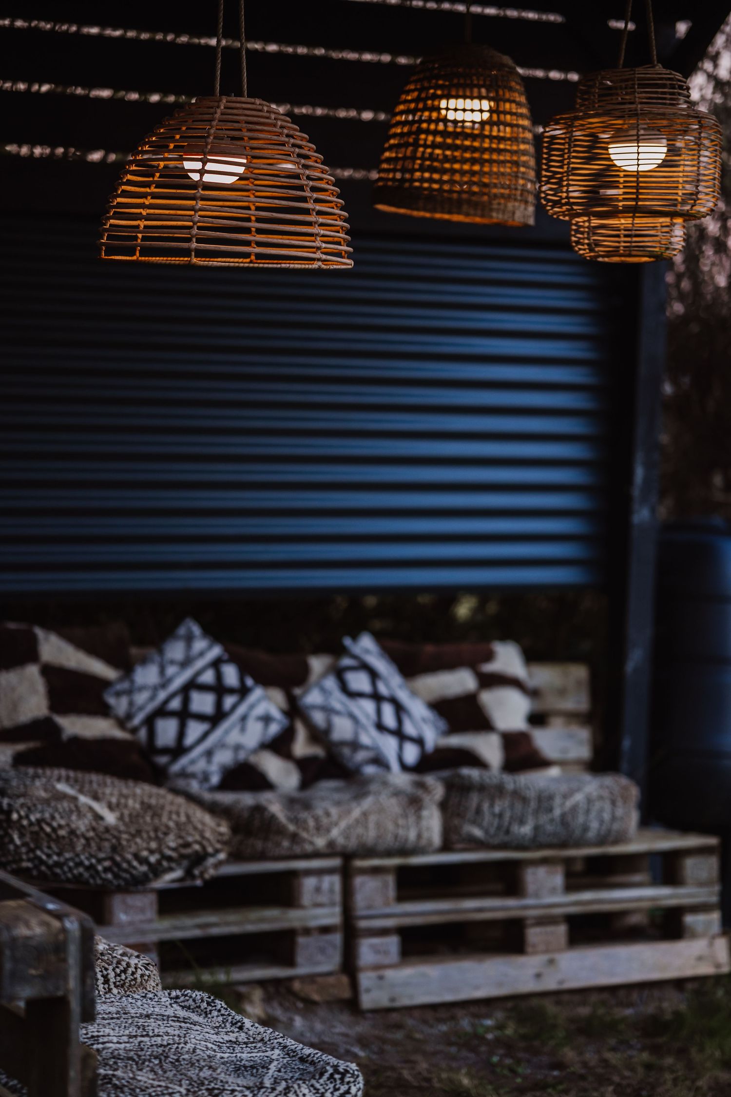 Rustic outdoor patio with wicker pendant lights illuminating pallet furniture with patterned cushions against a corrugated metal wall.