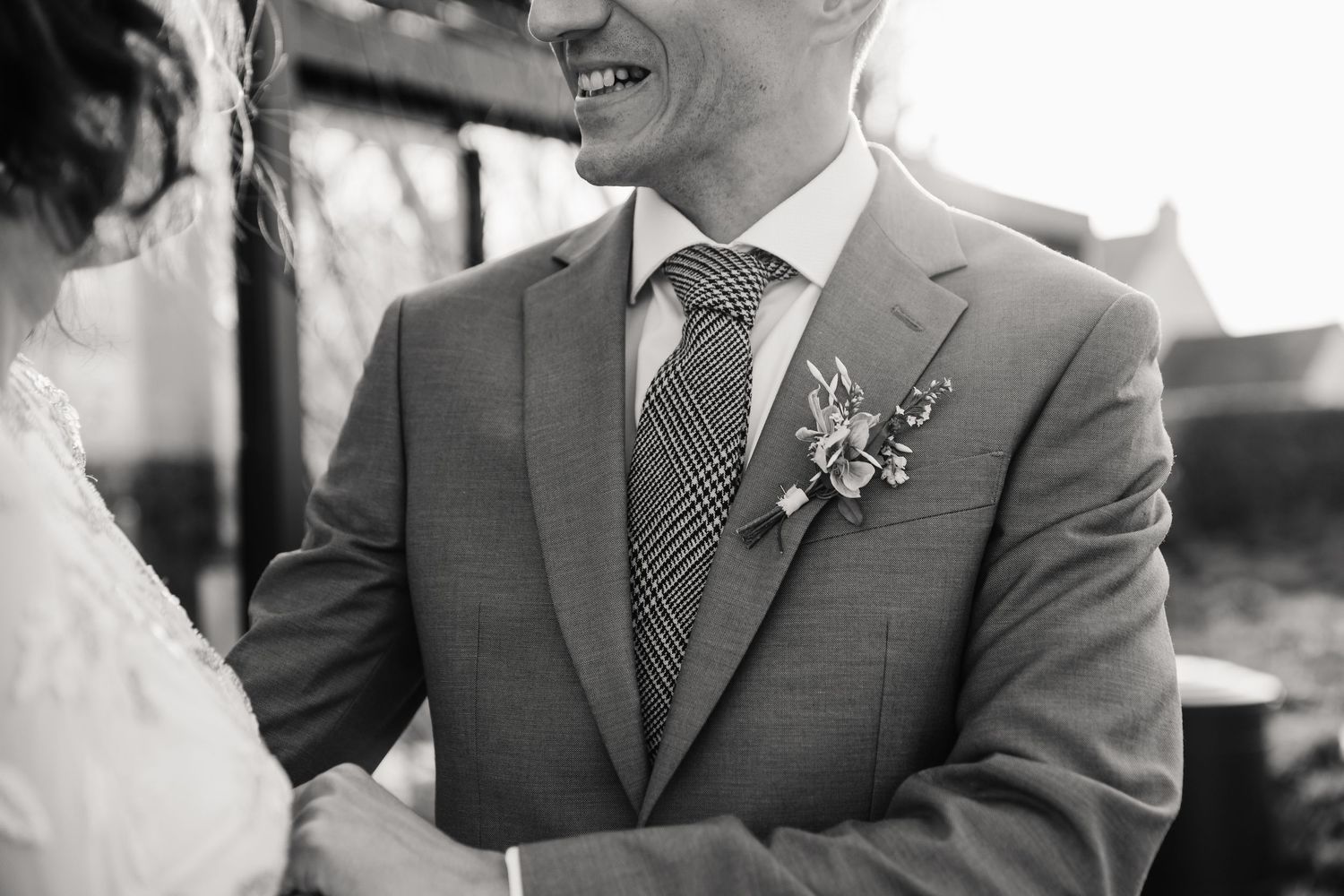 Black and white wedding photo of groom in gray suit with boutonniere and plaid tie, smiling at bride.