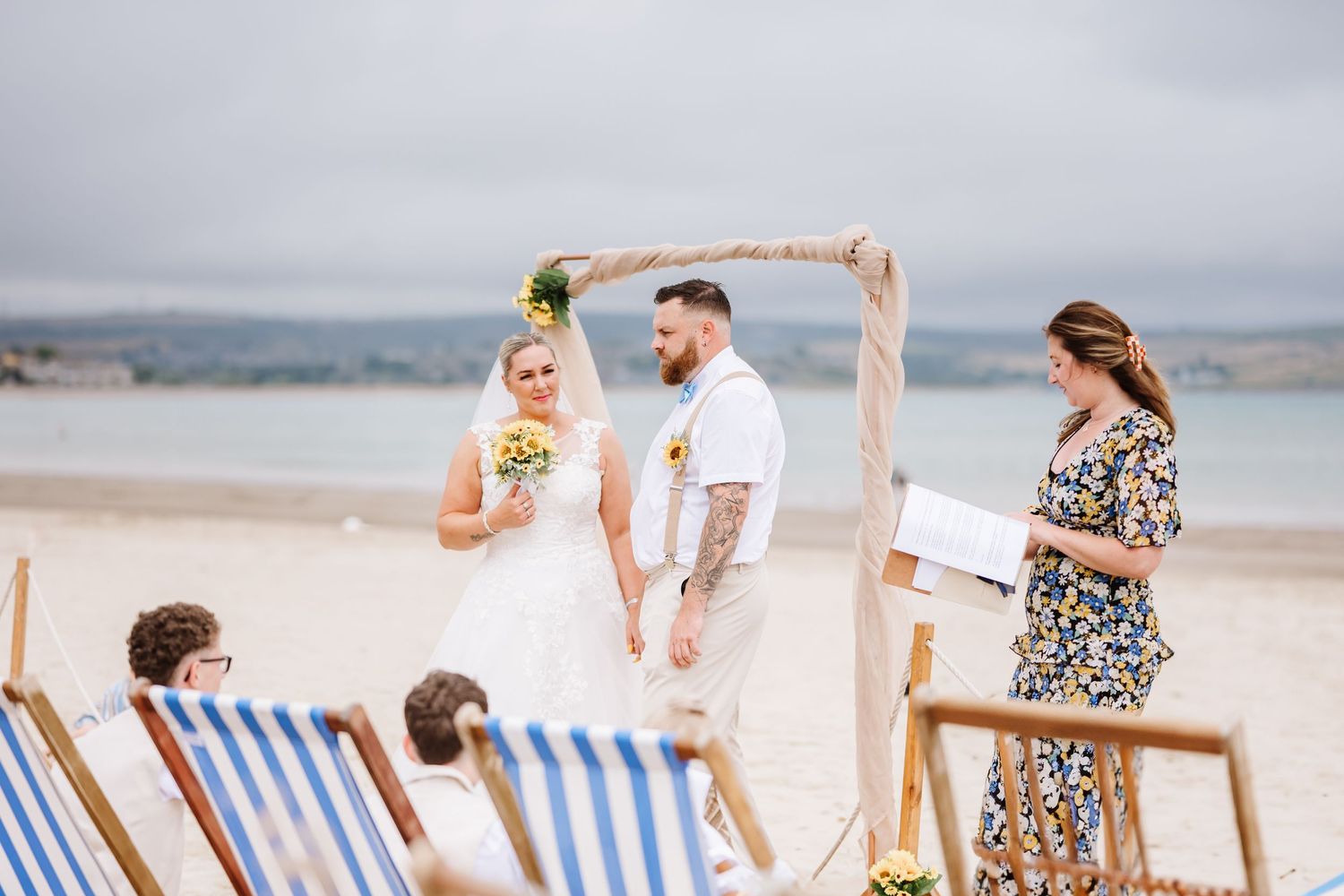 UK beach wedding on Weymouth Beach with Ceremonies by Laura Bell. Celebrant helps bride with dress. Sunflower wedding bouquet