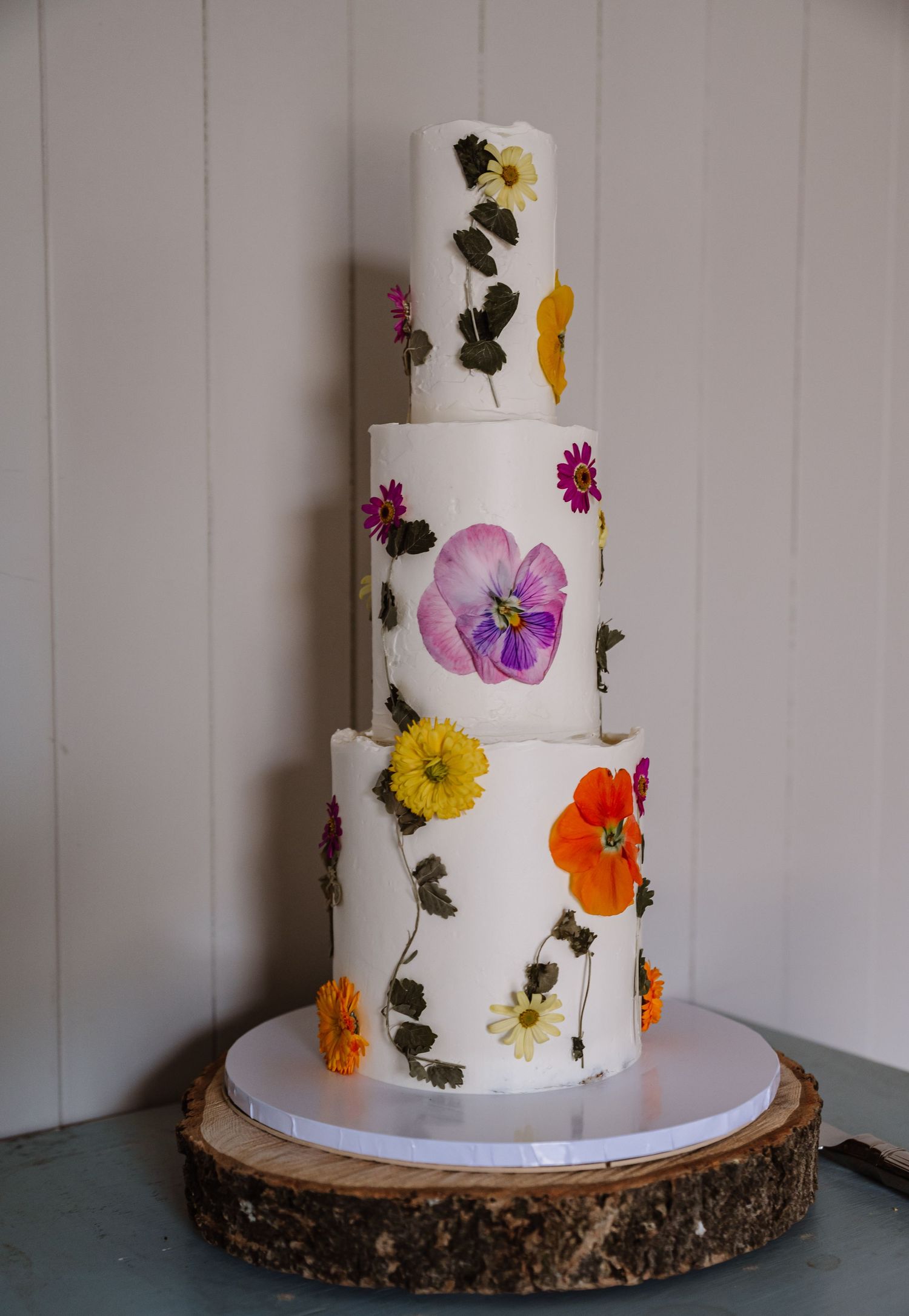 Four-tier white wedding cake decorated with colorful pressed flowers and leaves, displayed on a rustic wood slice stand. The Cornish Place