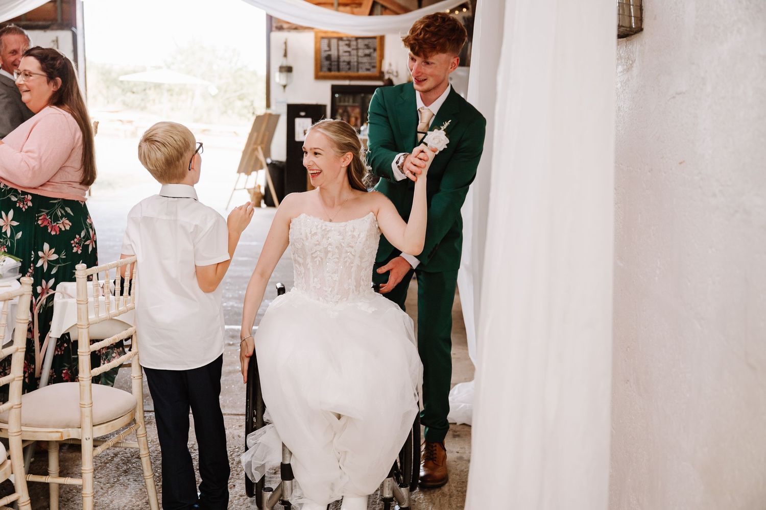 Smiling bride in a white lace wedding gown raises hands with a groom in a green suit while a young boy and guests watch indoors.