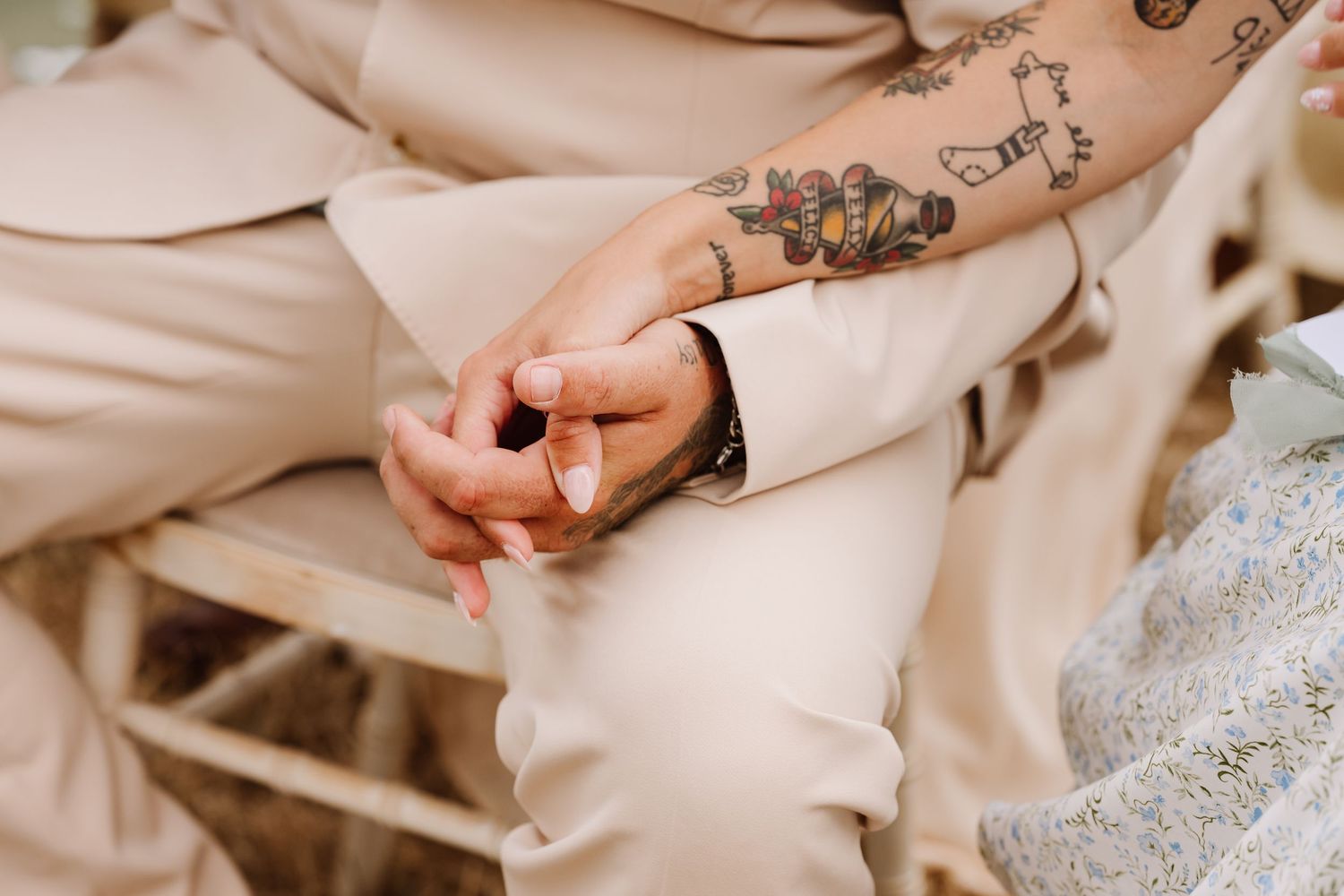 Close-up of couple holding hands, one with tattoos, wearing beige suits at a wedding ceremony.
