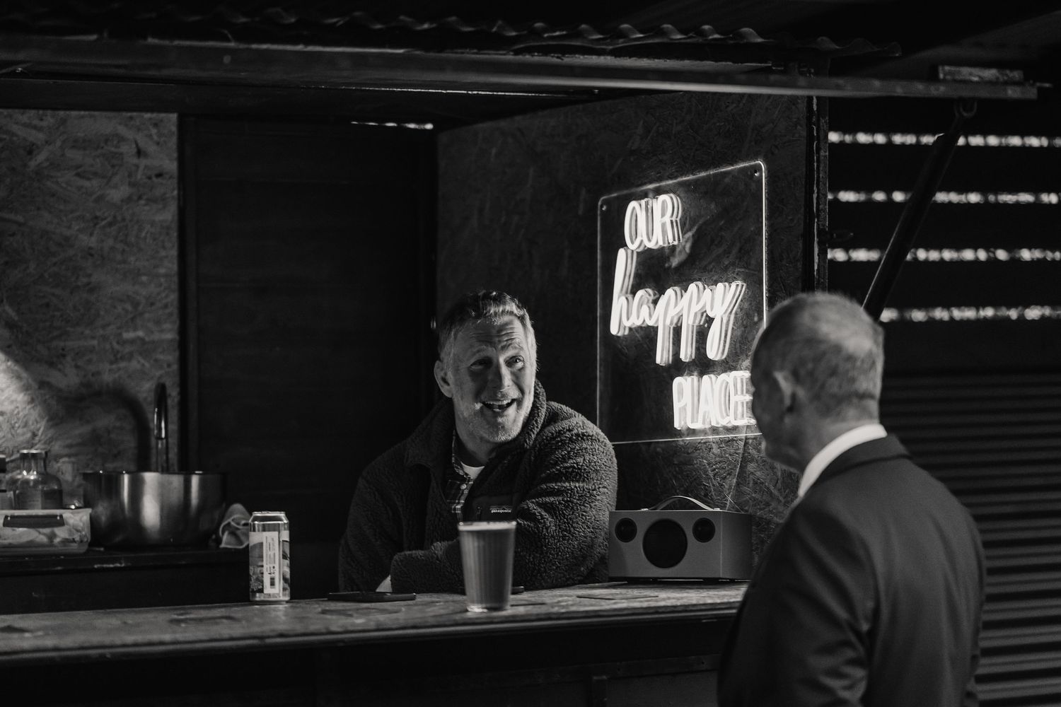 Black and white photo of two men conversing at a bar counter with a neon 'Our Happy Place' sign glowing behind. The Cornish Place,