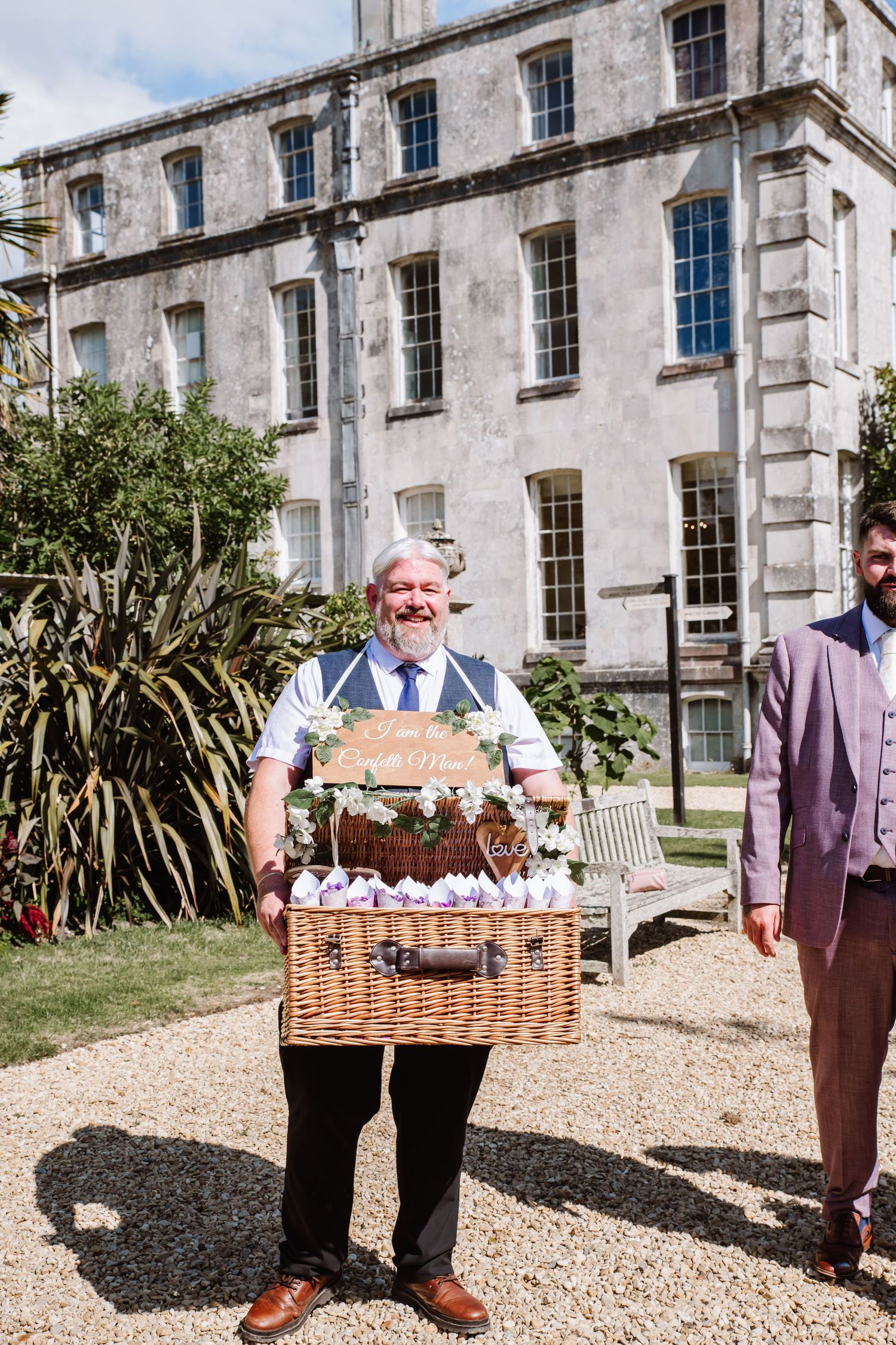 Chris Howat Dorset Magician holding a large wicker basket filled with items on a gravel path outside a historic building.