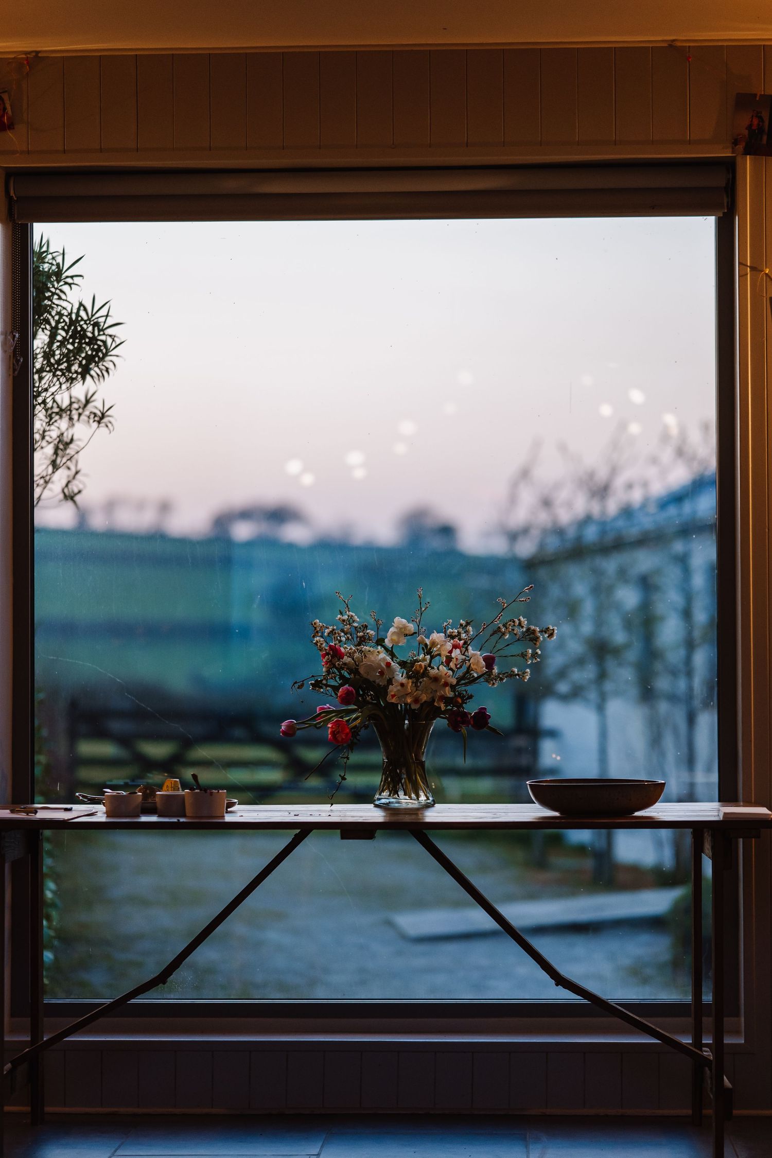 A floral arrangement in a glass vase on a console table beside a large window with a serene dusk garden view. The Cornish Place, wedding