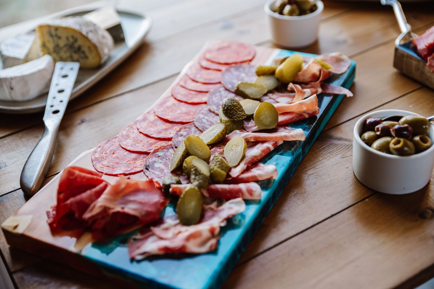 A charcuterie board with sliced salami, prosciutto, pickles, and olives on a wooden table.
