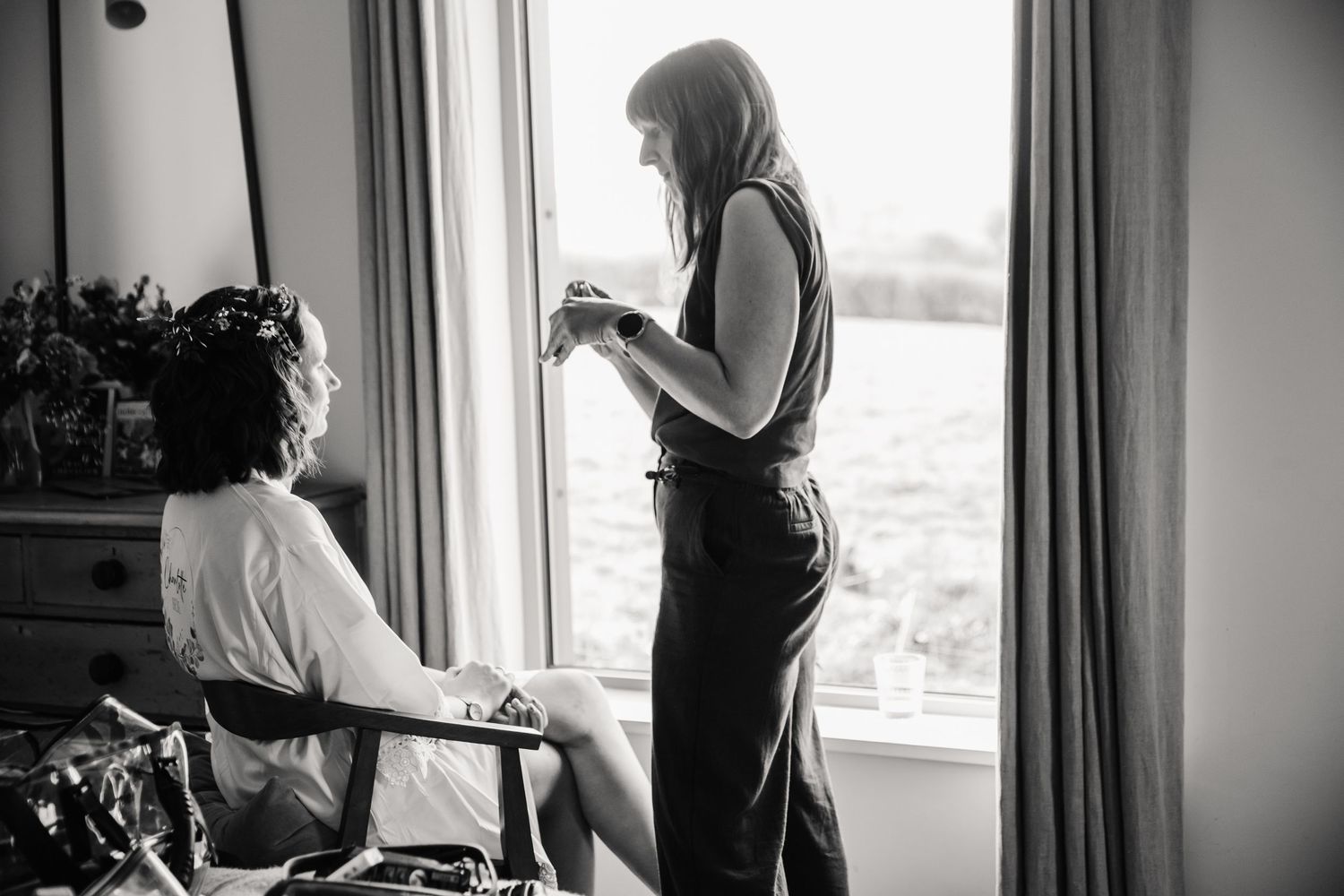 Black and white photo of a hairstylist styling a seated woman's hair beside a bright window while she gets ready for her wedding.