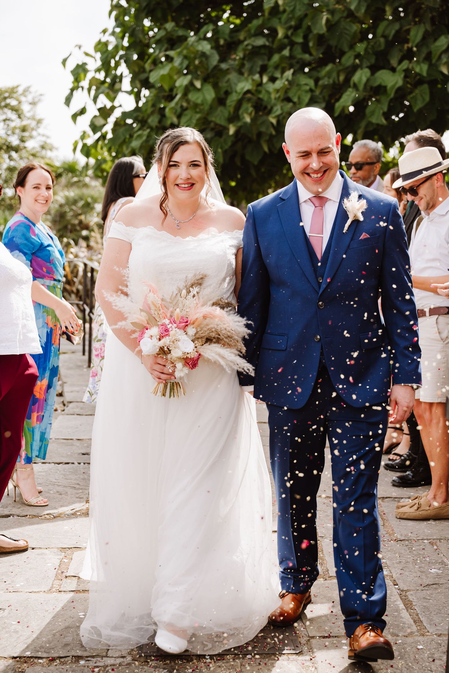 Bride and groom walk through confetti shower, she holds pink bouquet in white gown, he wears navy suit with pink tie at Westgate Southampton