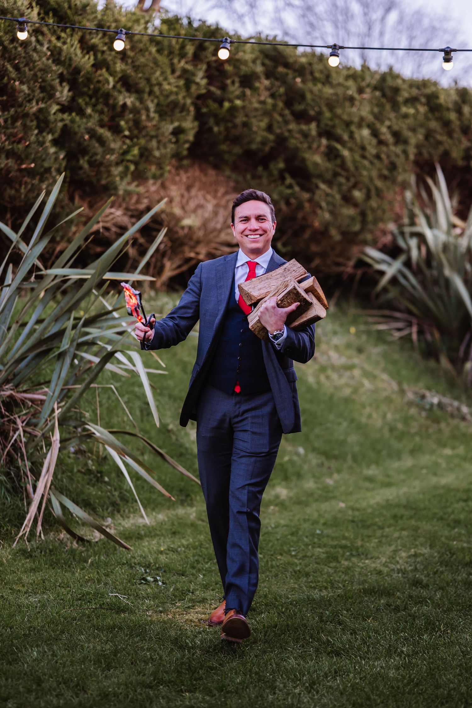 Smiling groom in navy suit carrying firewood and sparklers in a garden with string lights. The Cornish Place, Cornwall wedding venue