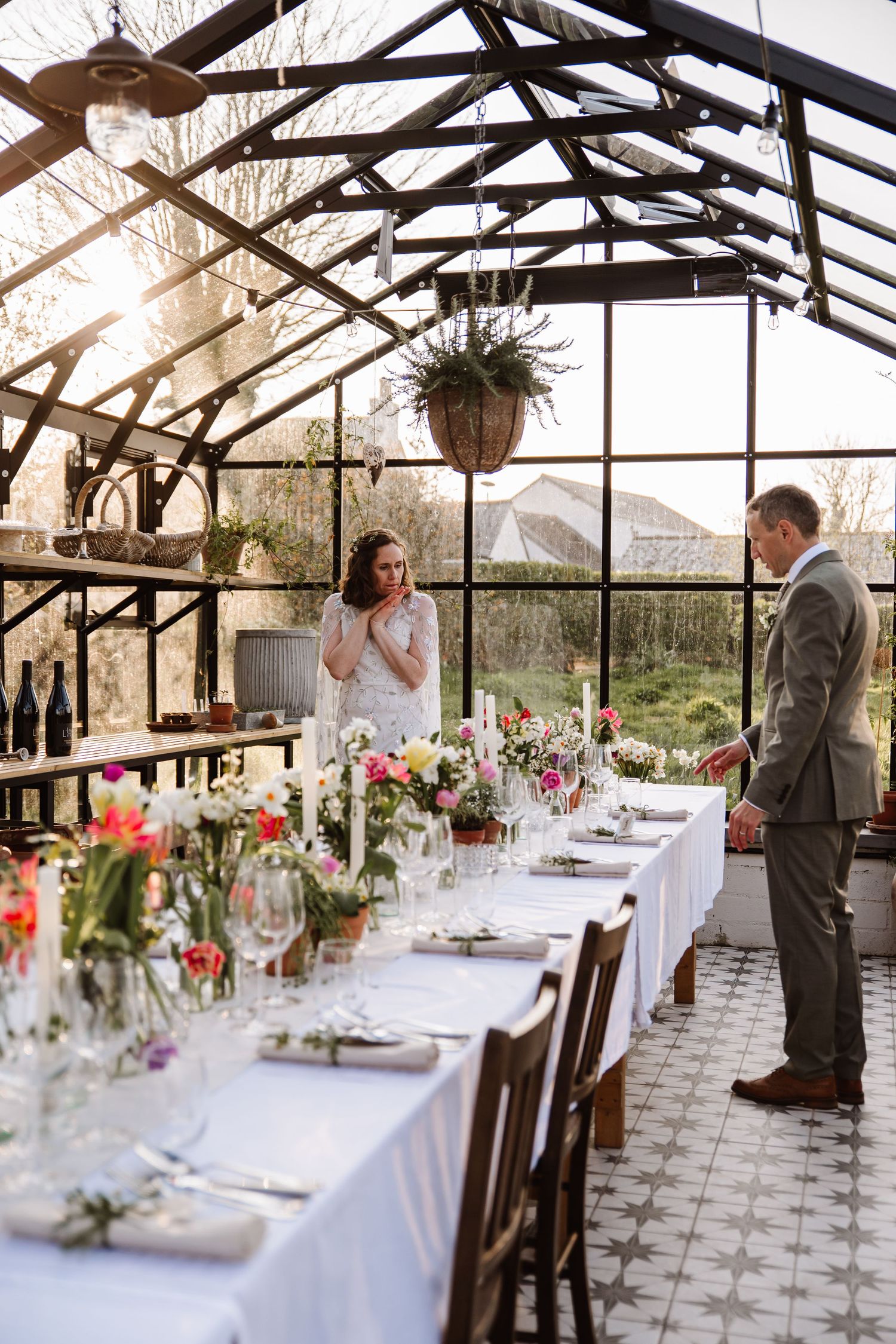 Bride and groom admire a floral-decorated wedding reception table inside a sunlit glass greenhouse venue. The Cornish Place wedding venue