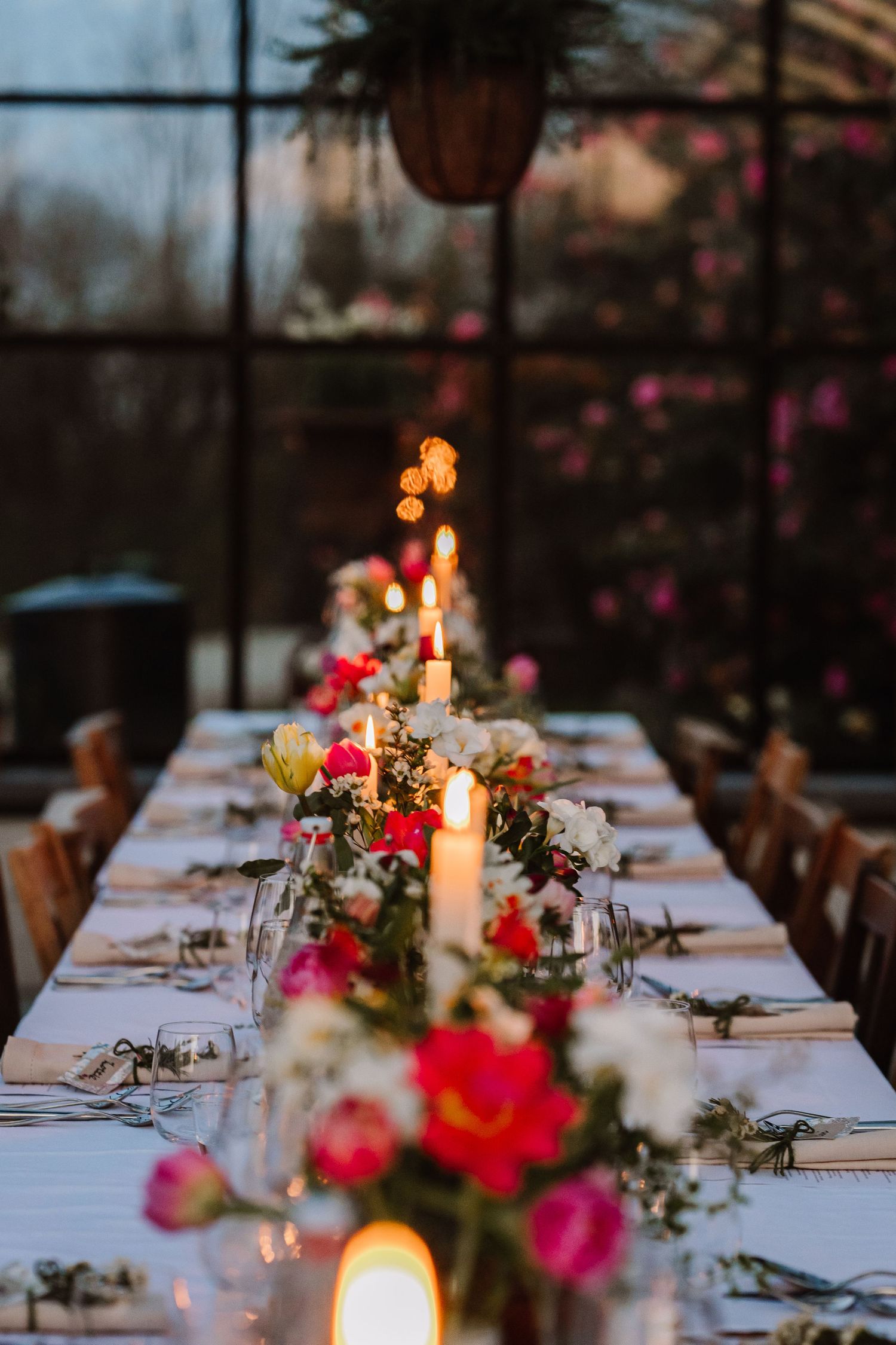 Pink blooming flowers frame an elegant glass greenhouse with long dining tables set up inside for an event. Unusual wedding venue cornwall