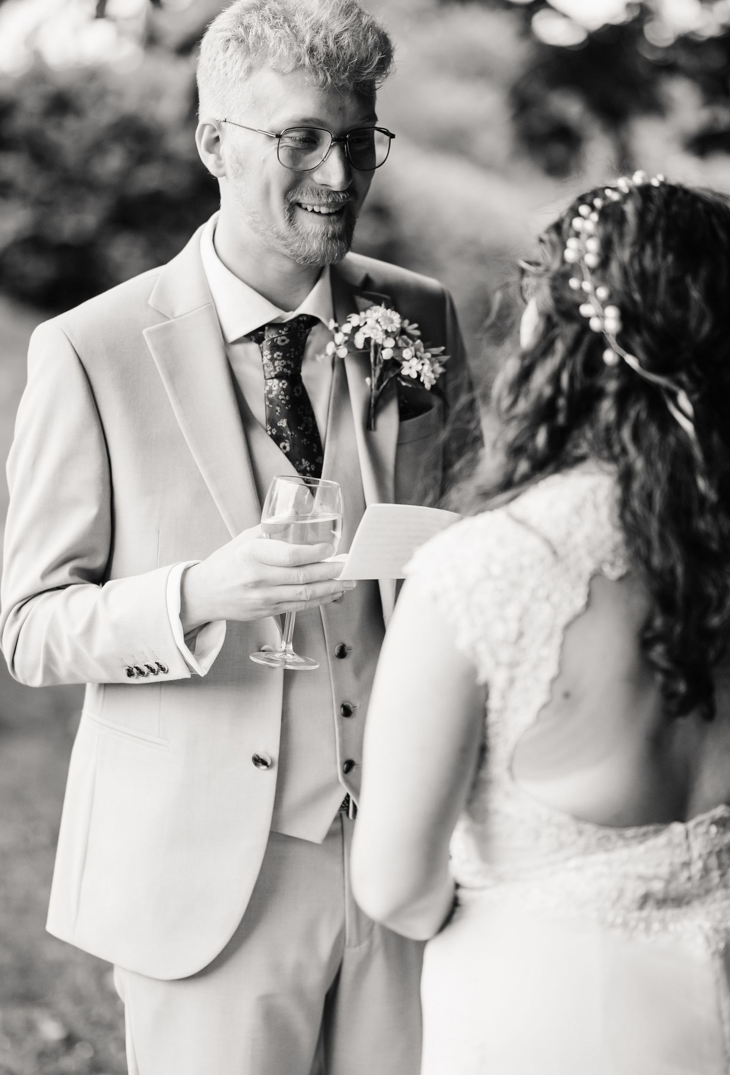 Black and white wedding photo of smiling groom in suit holding champagne, reading vows with bride in white dress at Symmondbury Tithe Barn,