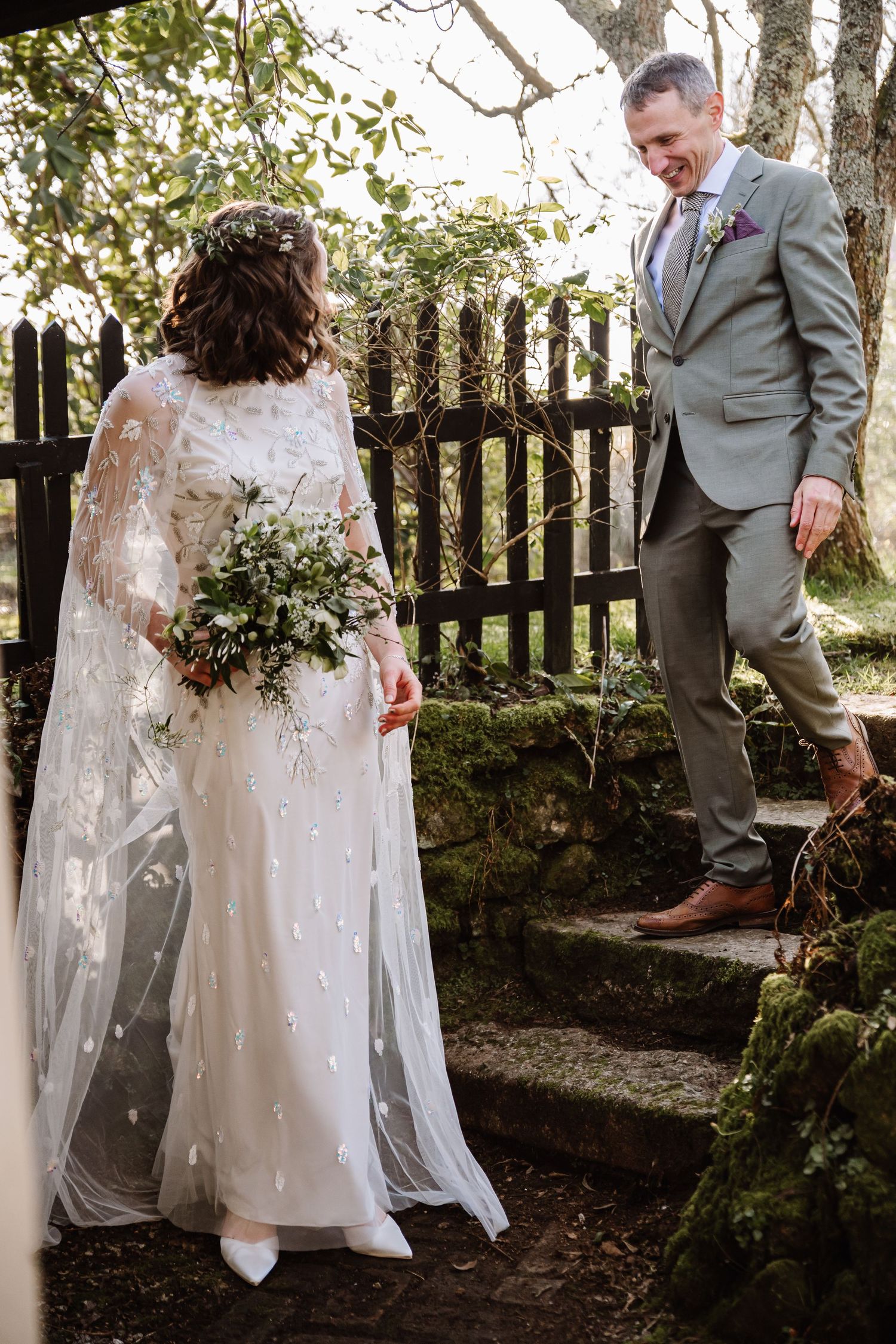 Bride in white gown with floral cape holds bouquet while groom in grey suit smiles on mossy stone steps outdoors at The Cornish Place