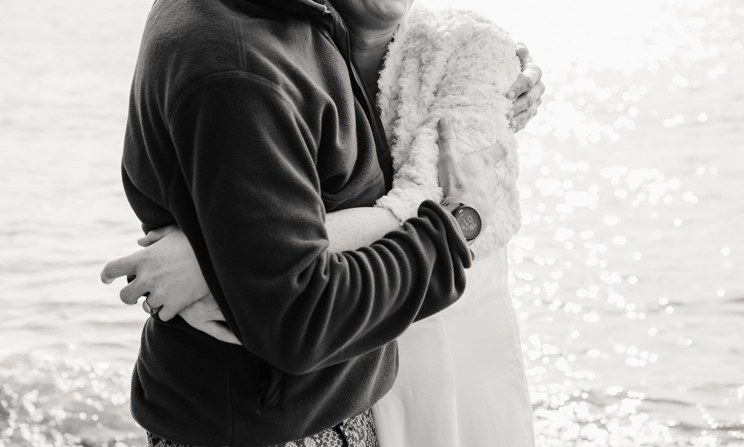 Black and white photo of a couple embracing romantically by the water, the woman wearing a white lace dress, Cornwall beach wedding day