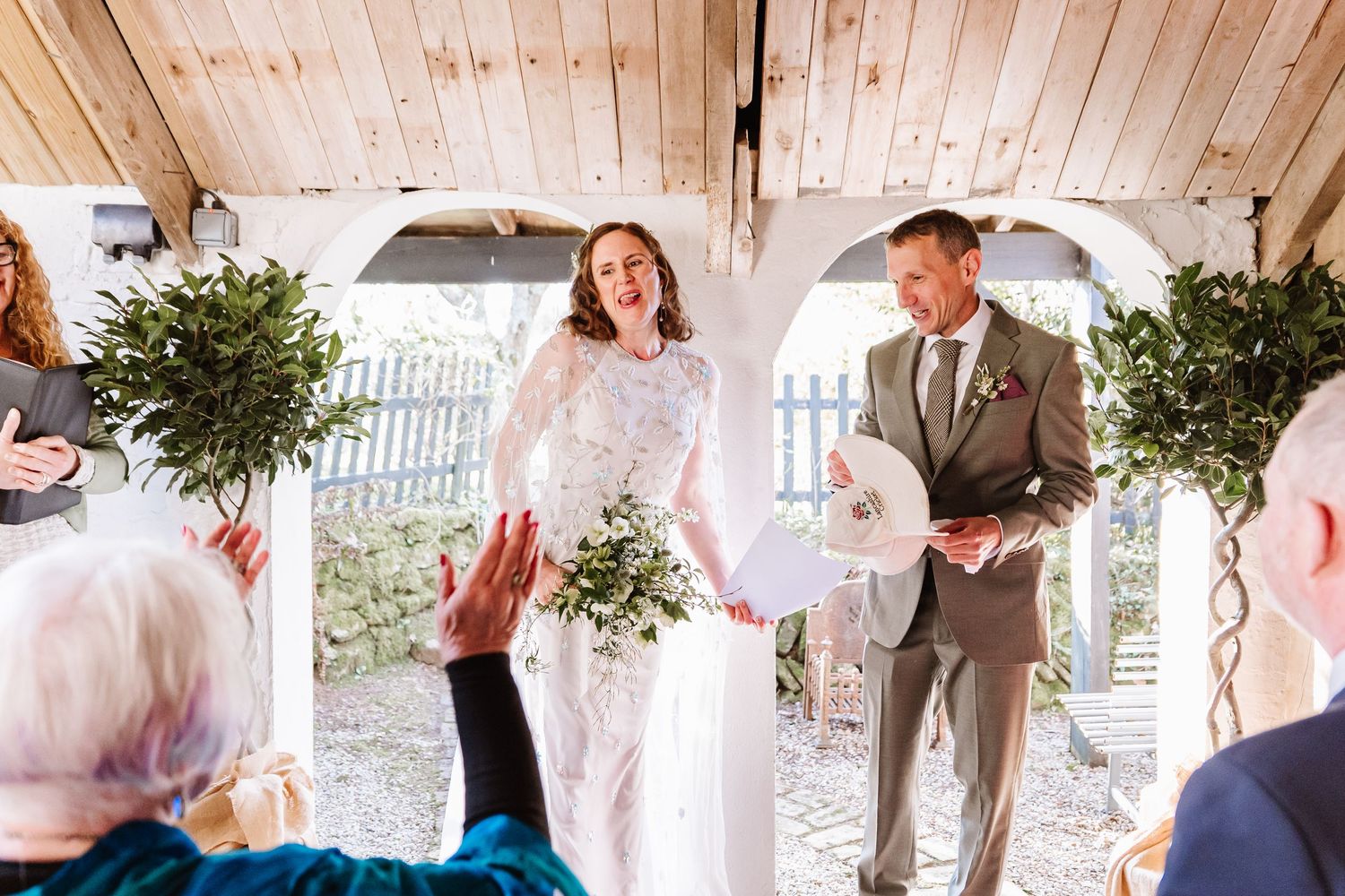 Bride and groom laughing joyfully during outdoor wedding ceremony beneath white arch with greenery. The Cornish Place, Wood shed wedding
