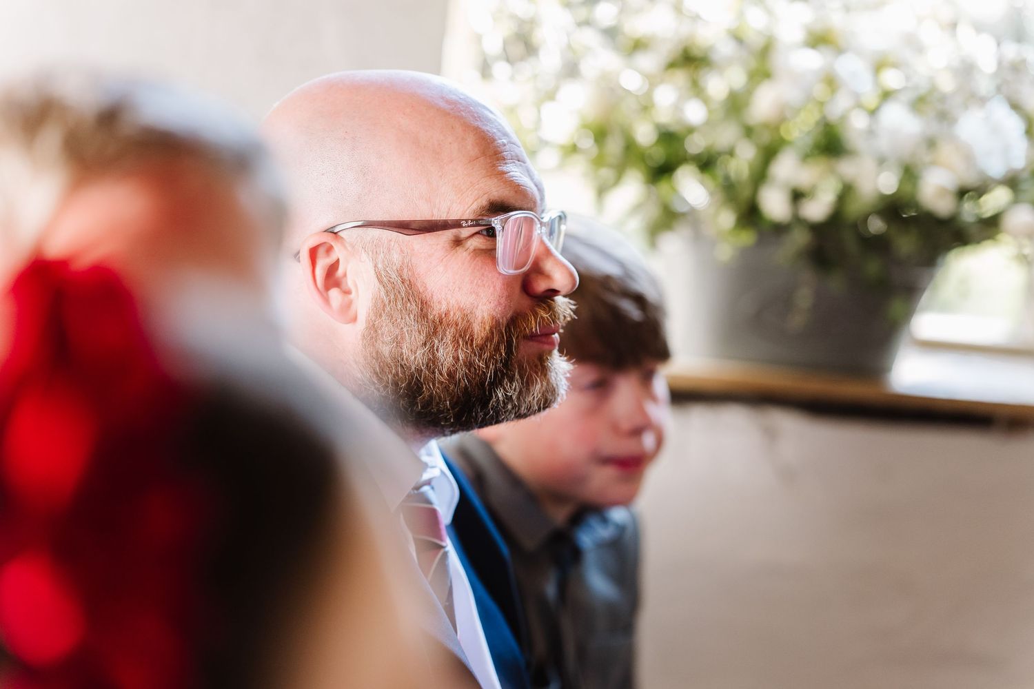 Bald bearded man with glasses sitting thoughtfully at an indoor event with blurred figures and flowers in background.