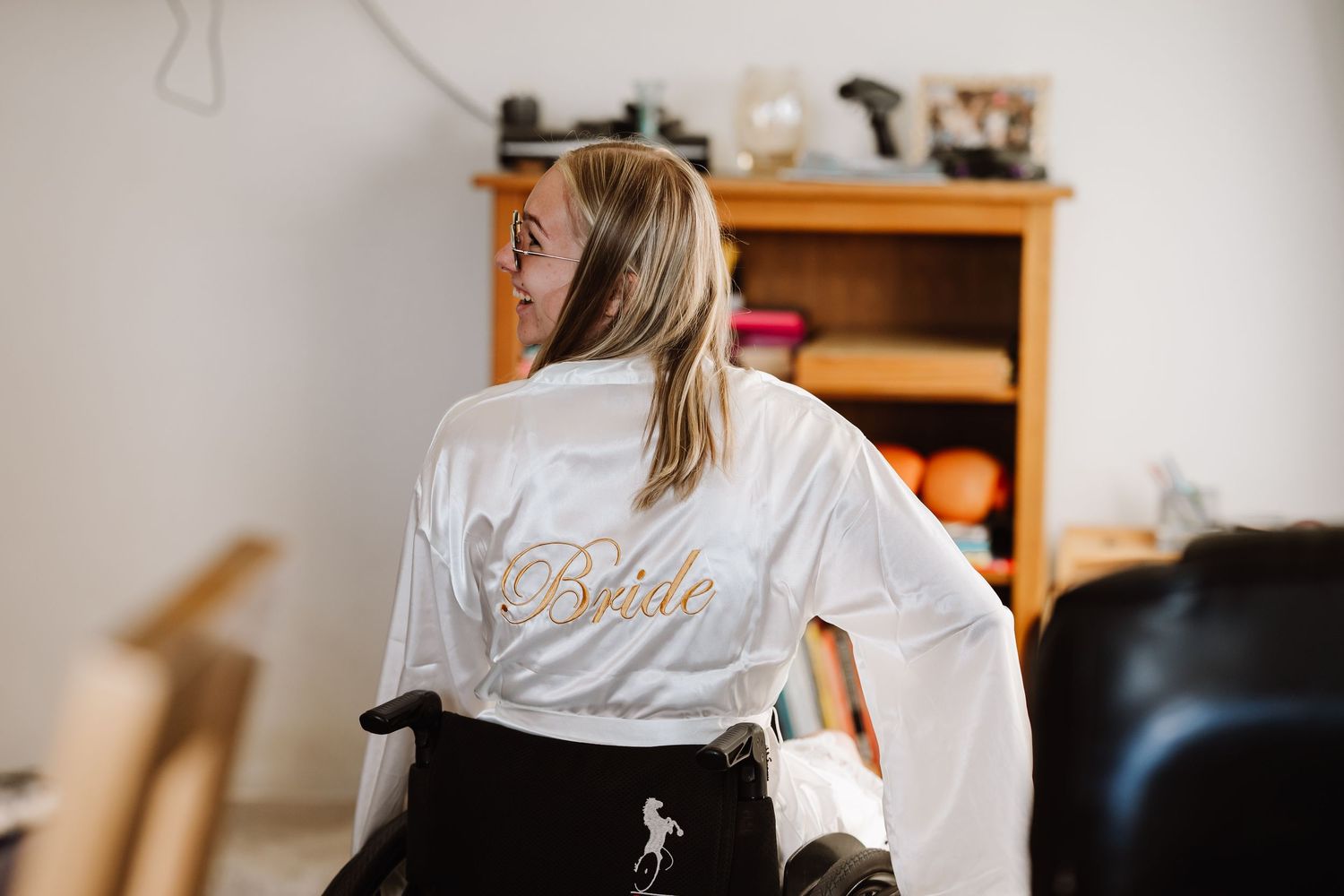Bride in wheel chair wearing a white satin robe with gold script lettering, standing in a bright room before her wedding.