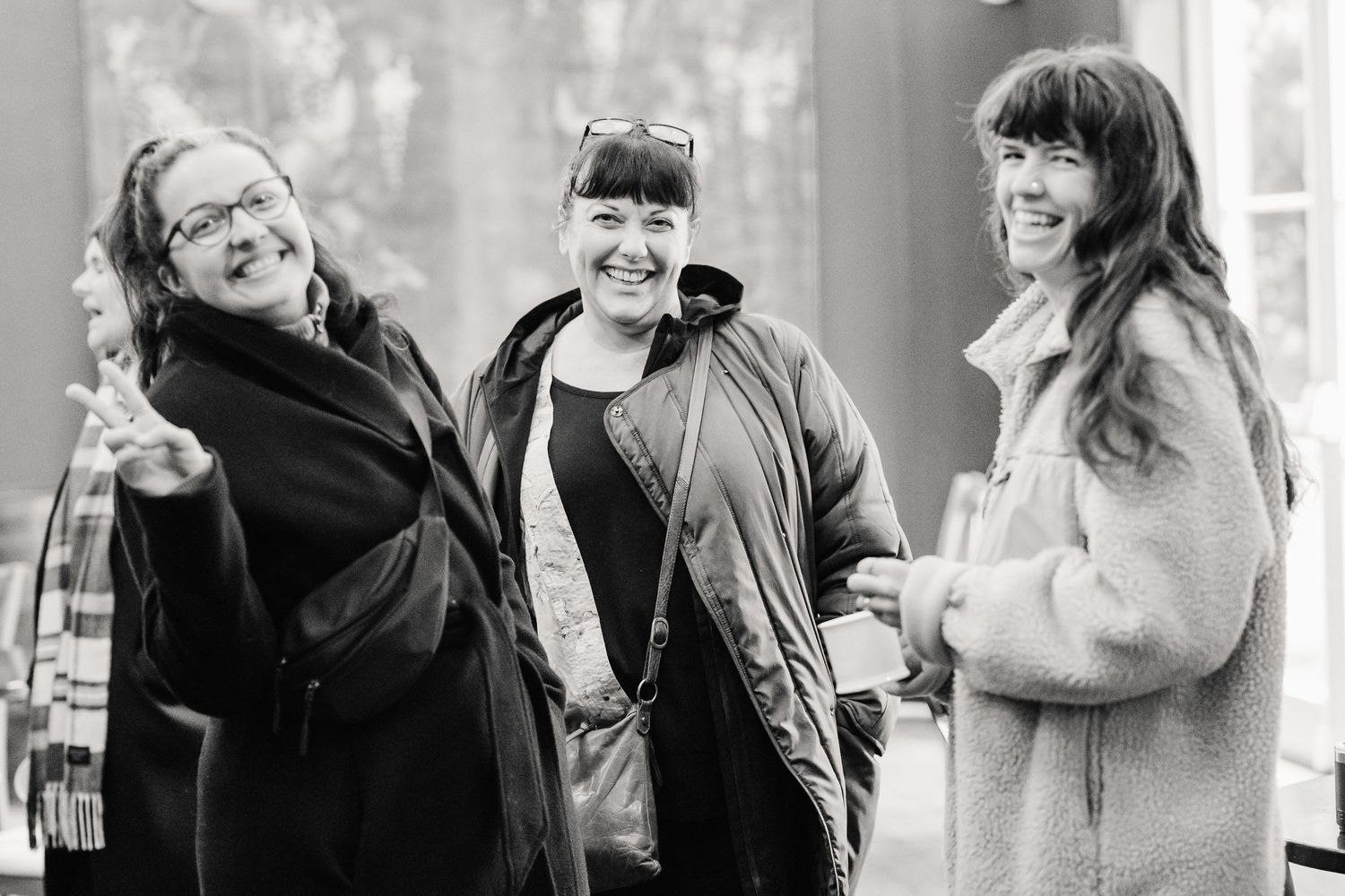 Three smiling women in coats laugh and chat together indoors in a joyful black and white photo, one waving at the camera.