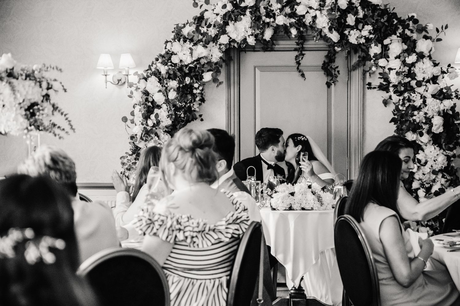 Black and white wedding reception photo of couple kissing at sweetheart table beneath floral arch Wedding at Hotel Du Vin Winchester