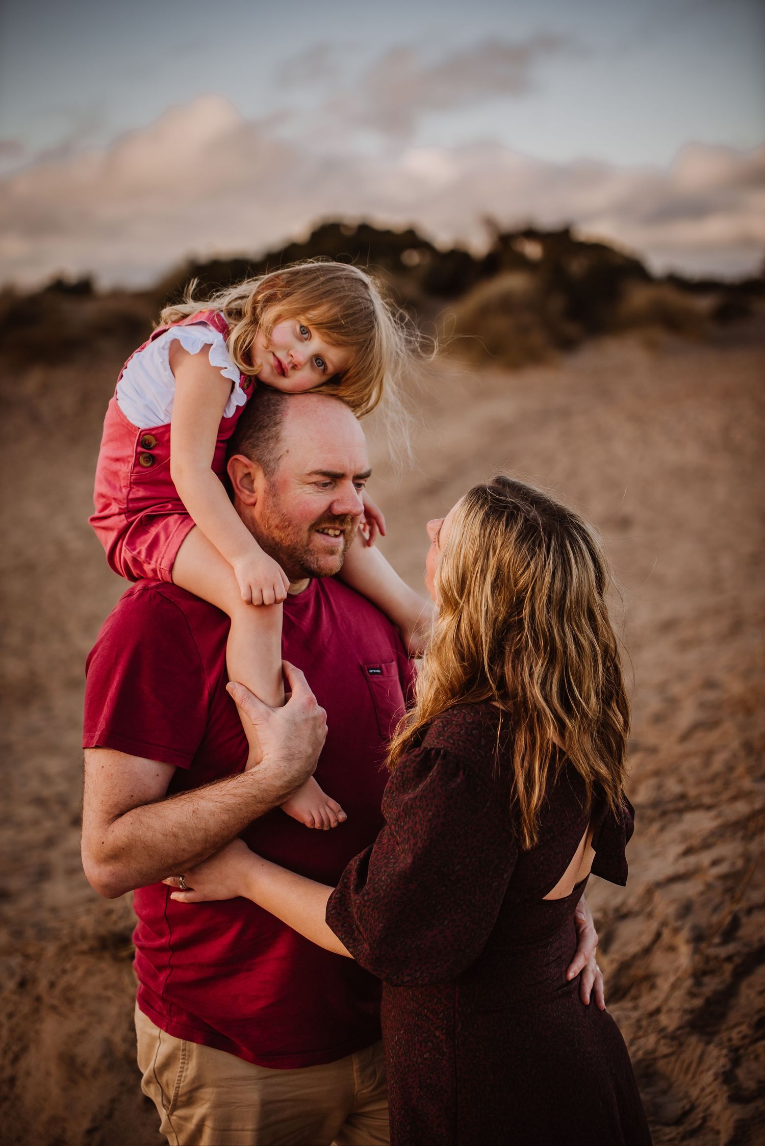 A joyful family moment on a sandy beach, with a little girl sitting on her fathers shoulders while mom smiles up at them.