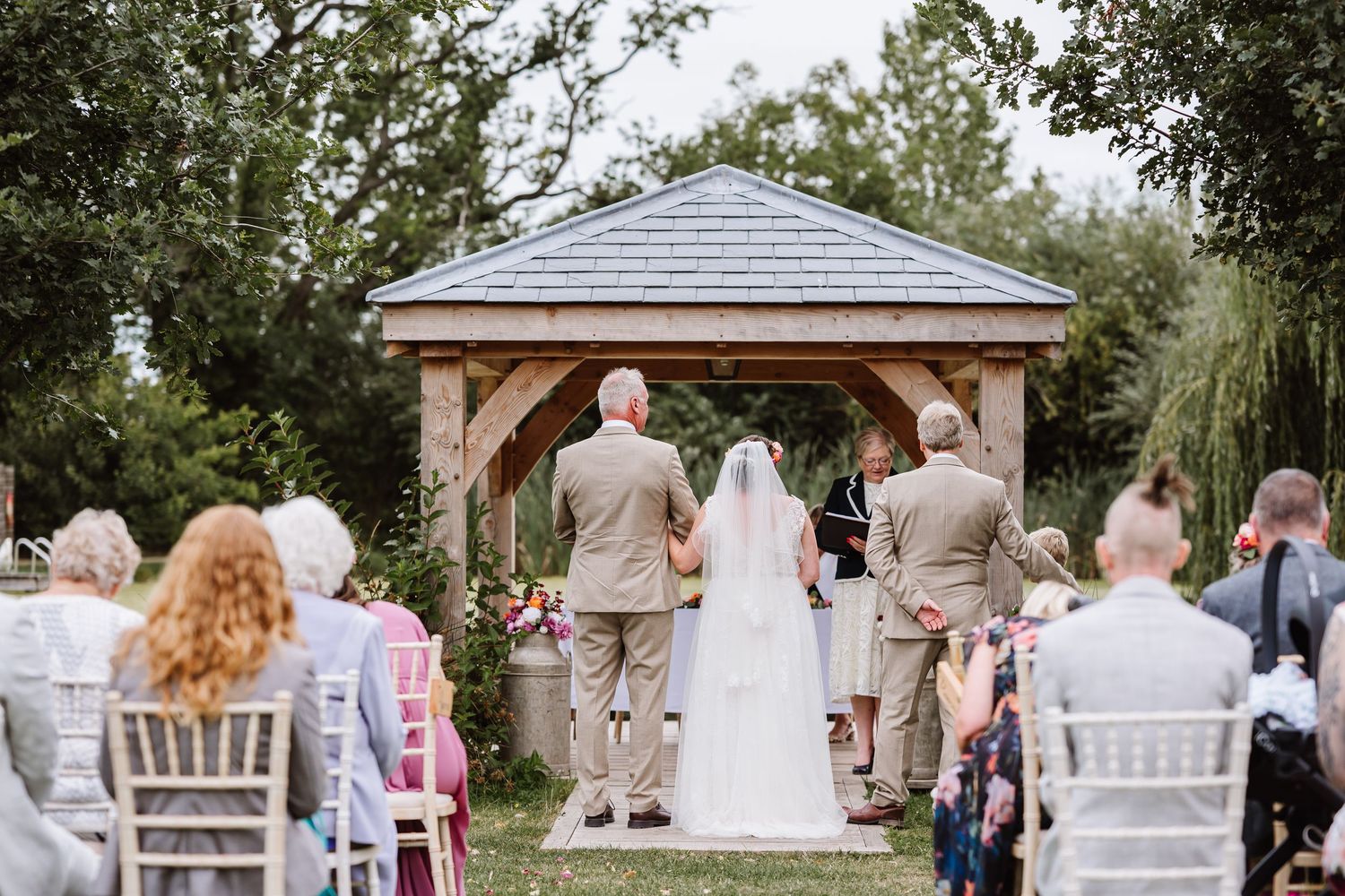 Wedding ceremony guests gather under trees and a wooden gazebo during an outdoor ceremony at Waterside Country Barn and Mill