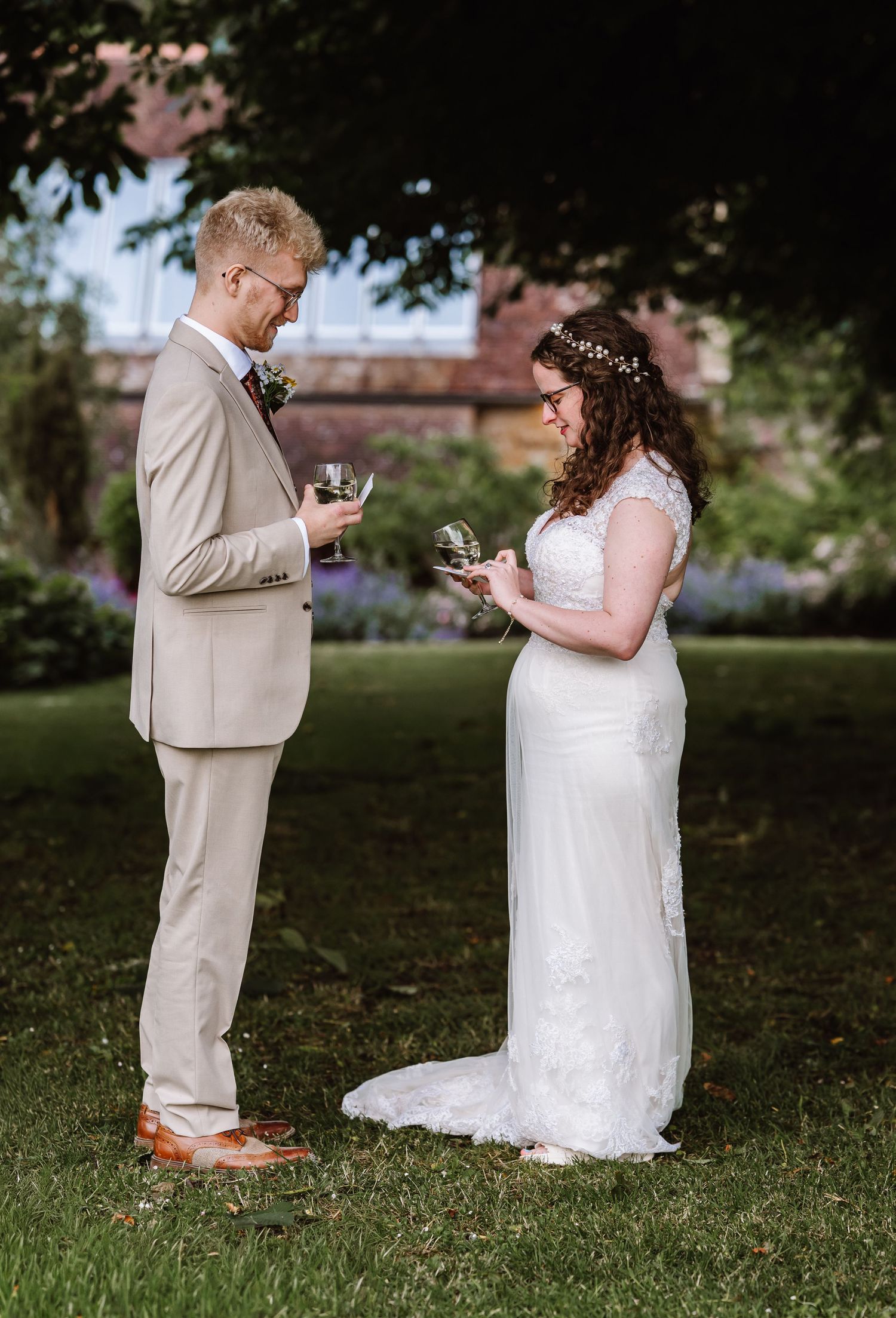 Bride and groom exchange rings outdoors in garden, she wears lace dress with floral crown, he wears tan suit at Symmondbury Tithe Barn, Brid
