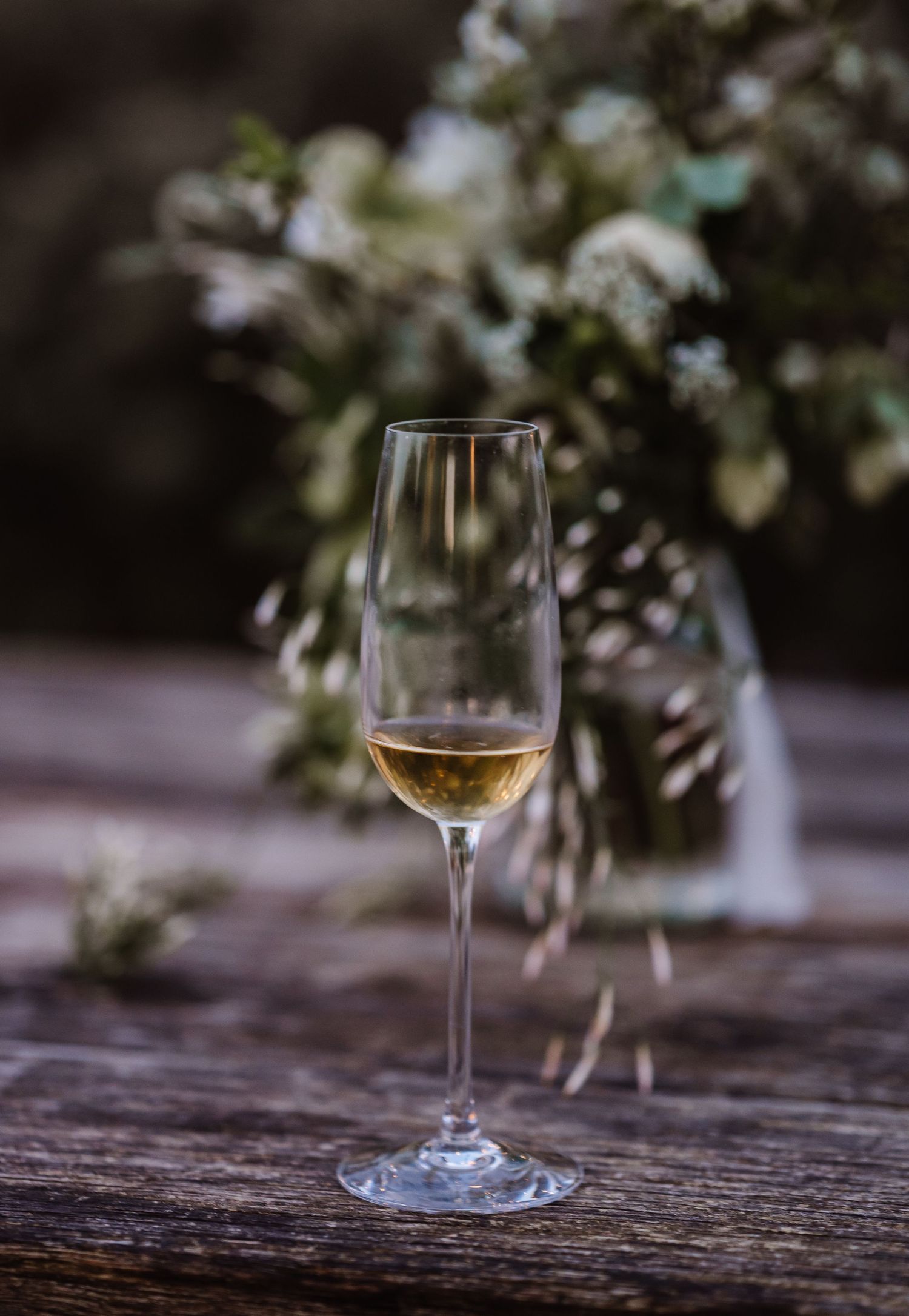 A delicate champagne flute filled with white wine on a rustic wooden table with a floral arrangement in the background.