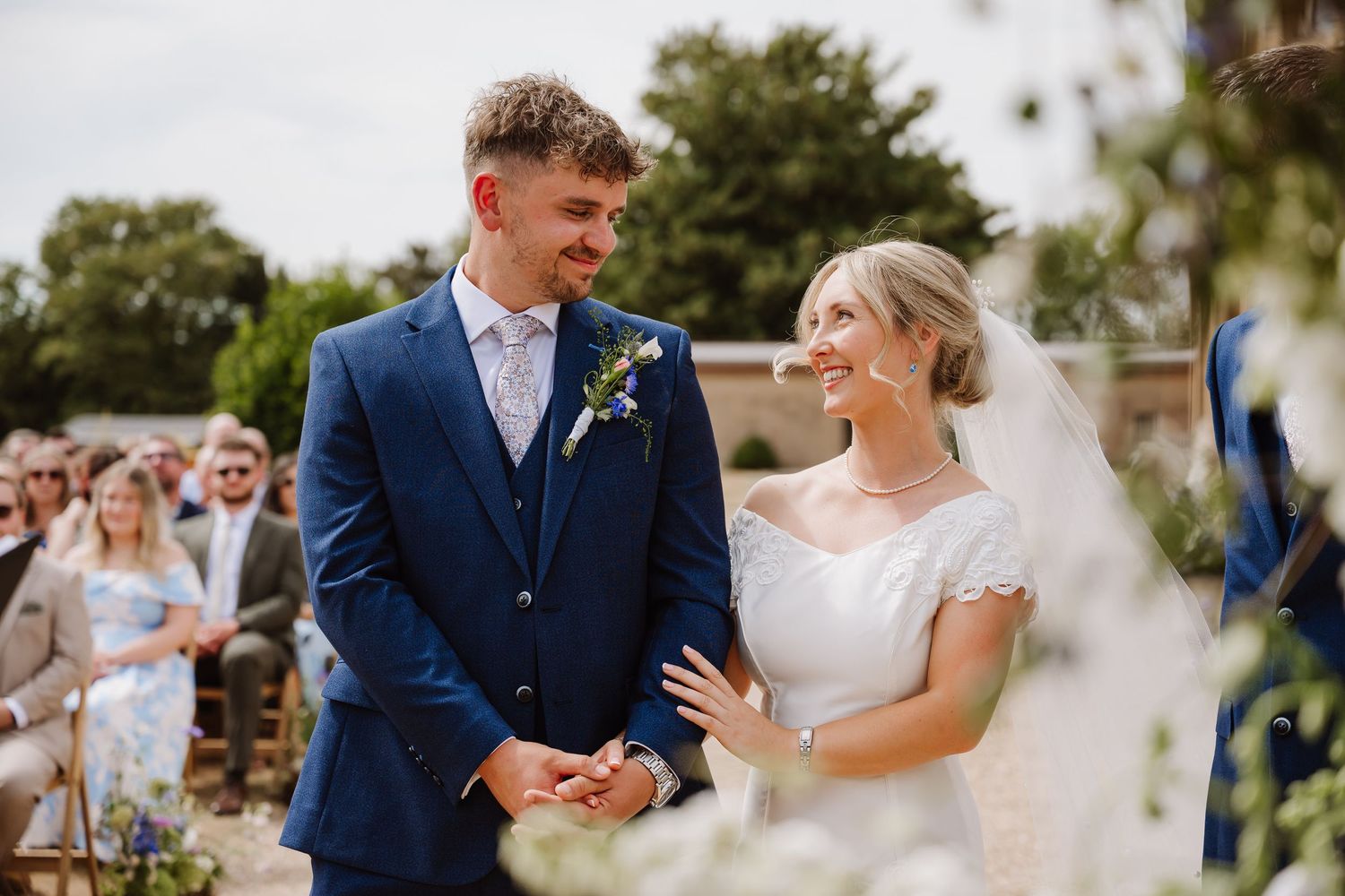 Bride and groom smiling at each other during outdoor wedding ceremony, groom in blue suit, bride in white dress.