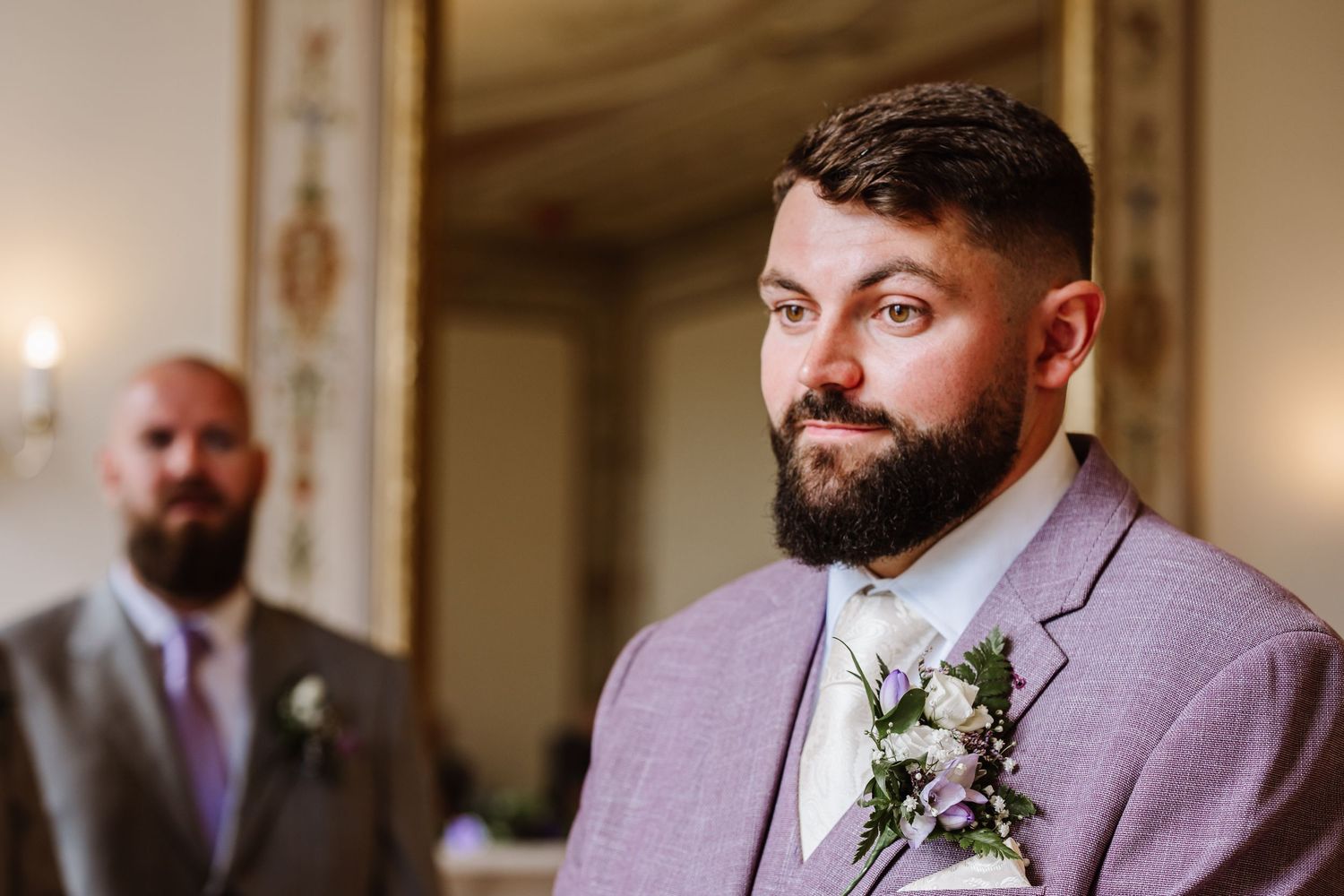 A groom wearing a light purple suit jacket with a floral boutonniere poses in a warmly lit indoor setting. at Kingston Maurward Estate
