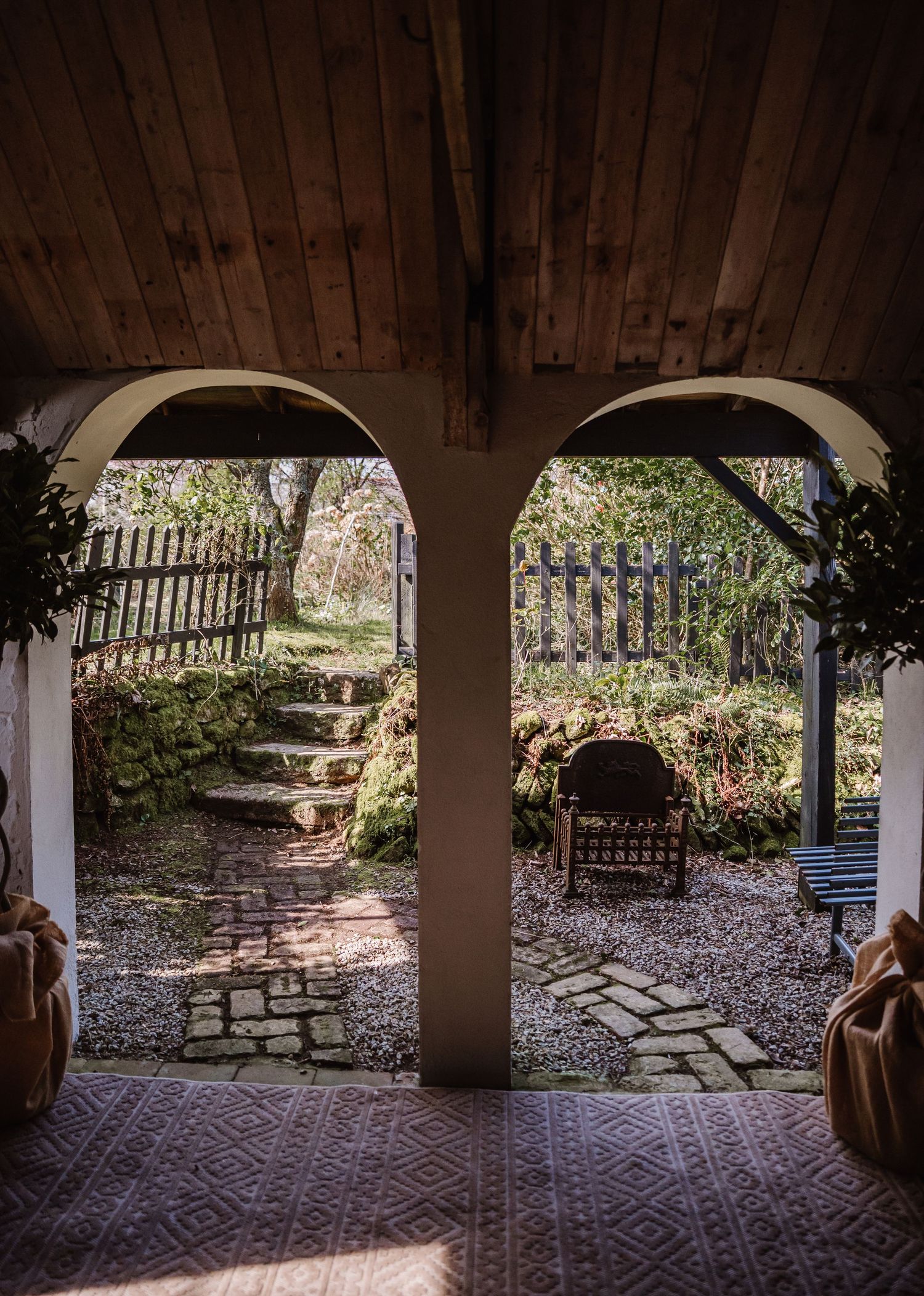 Rustic covered porch with stone arches overlooking a charming garden with cobblestone path, wooden fence, and lush greenery.
