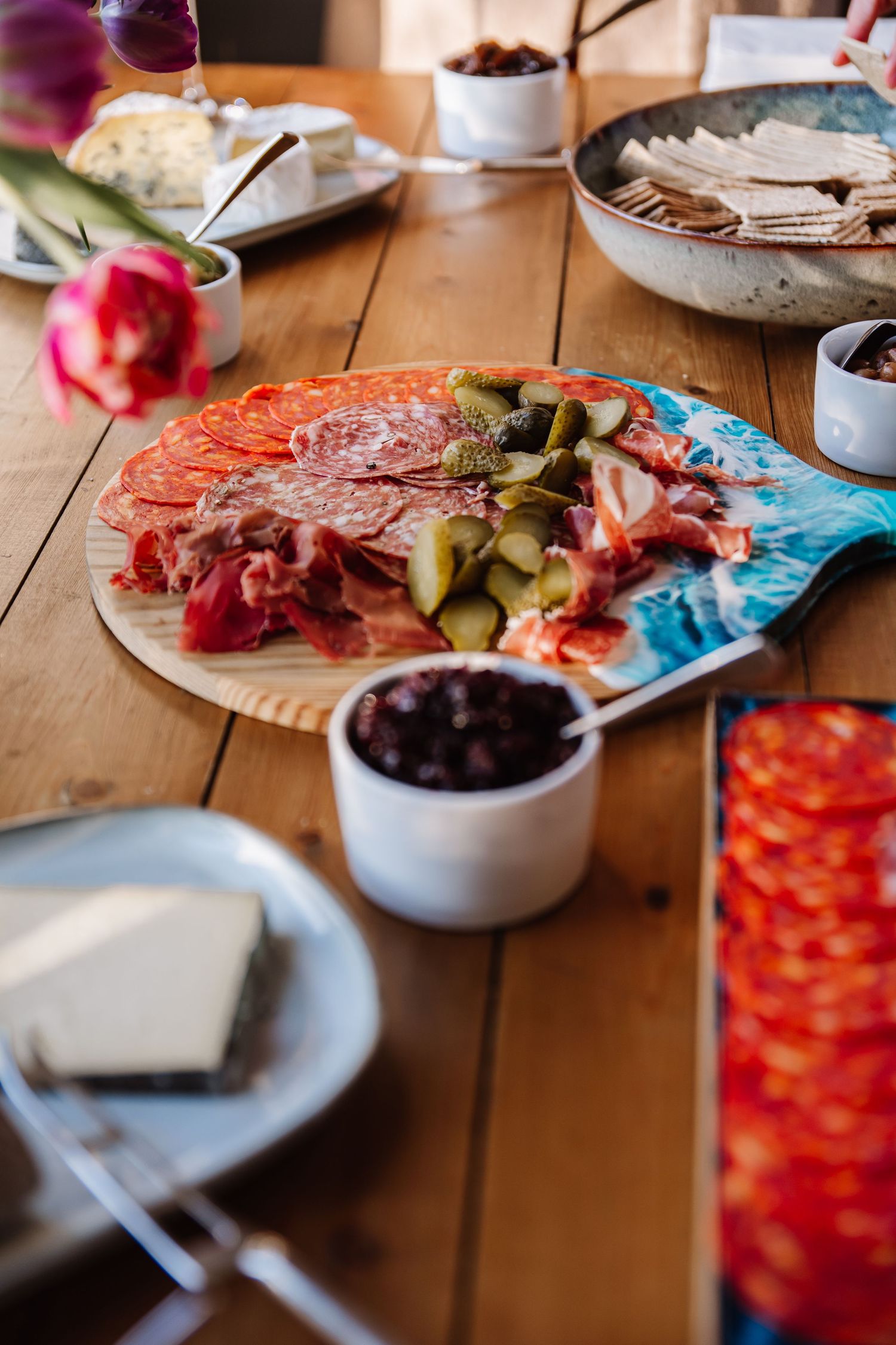 A rustic wooden table with charcuterie board featuring cured meats, pickles, olives, cheese, and crackers with a pink tulip. Cornwall weddin